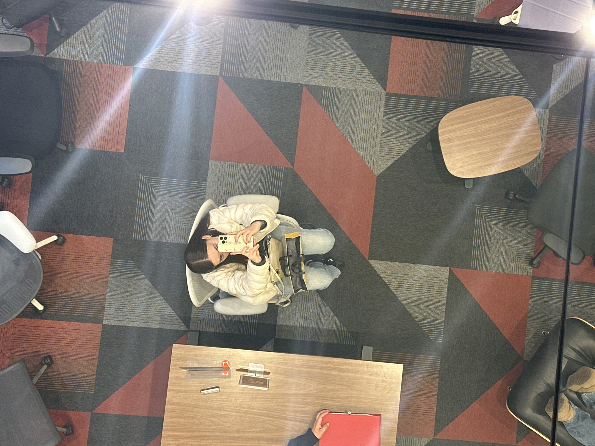 The image presents an overhead view of an interior space, possibly an office or co-working area, in Mexico City, Mexico. The floor is covered with a patterned carpet featuring geometric shapes in red, grey, and black. Several pieces of furniture are visible, including office chairs of various styles, a round wooden table, and a rectangular wooden desk. A person is seated in a chair, wearing a white jacket and light blue jeans. They are holding a mobile phone and appear to be taking a picture. Another person's hand is visible at the bottom of the frame, interacting with a red folder on the desk. Various small objects are present on the desk, suggesting a workspace. The overall impression is a modern, functional workspace.
