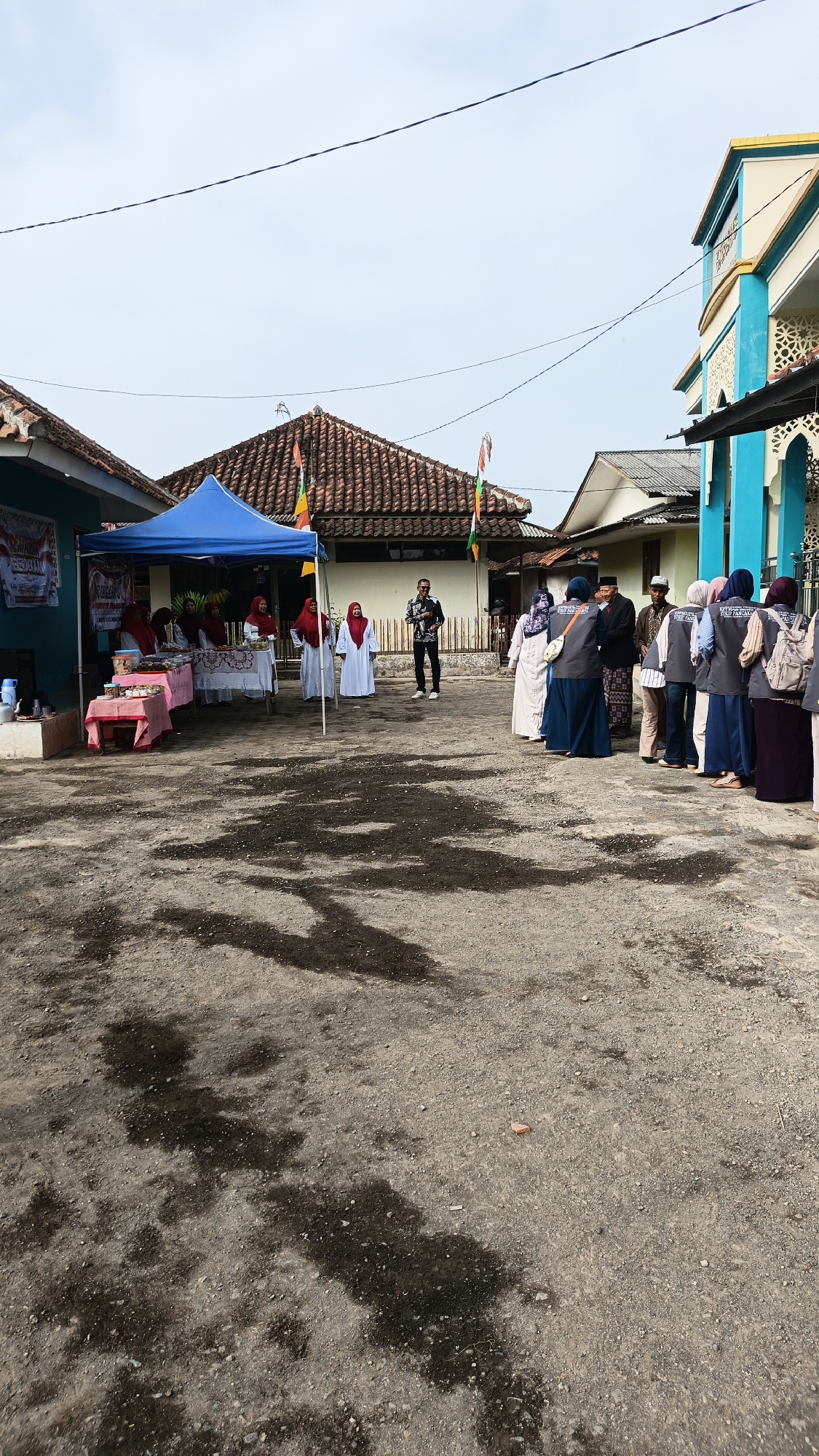 In Linggasirna, Indonesia, a group of people are gathered outdoors in front of several buildings. The ground is a mixture of dirt and gravel with some dark, wet patches. To the left, a blue tent is set up over a table laden with food items. Several women in hijabs are attending the table. A man in a patterned shirt and dark pants stands nearby, looking towards the right. A line of people, mostly women in hijabs and vests, are standing on the right side of the scene, facing a building that appears to be a mosque with turquoise and yellow accents. One woman in the line is wearing a light-colored hijab and a white traditional dress. Two women in the foreground on the right are wearing light-colored hijabs and dark blue dresses. Another woman in the foreground on the far right is wearing a purple hijab and a dark top. The sky is overcast with some thin clouds. Decorative flags are visible on poles in the background. The overall scene suggests a community event or gathering.