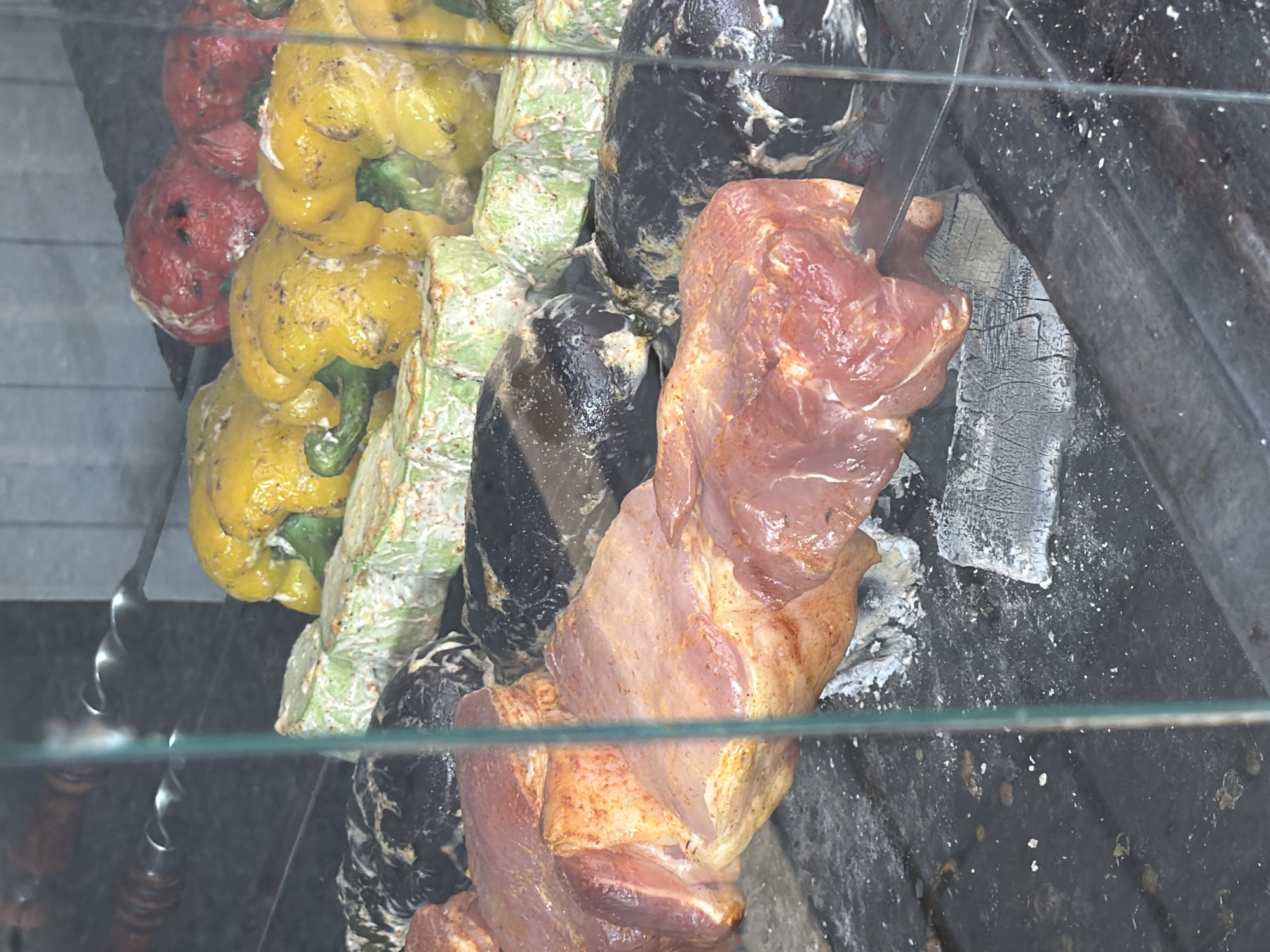 This media file shows skewers of meat and vegetables being grilled over hot coals. The subjects include pieces of raw, seasoned meat and what appear to be bell peppers and possibly eggplant. The peppers are yellow and red, and some have green stems. The vegetables are interspersed with the meat on the skewers. The scene is set in an outdoor or enclosed grilling area, with visible charcoal embers and grey ash. A metal skewer is partially visible, with a spiral design. A knife is also present, appearing to be used to prepare or adjust the meat on the skewer. The lighting suggests it is daytime. There is a glass barrier in the foreground, reflecting some of the scene. The location is identified as Sochi, Russia. No people are visible in the frame. The overall impression is that of food preparation, likely for a meal.