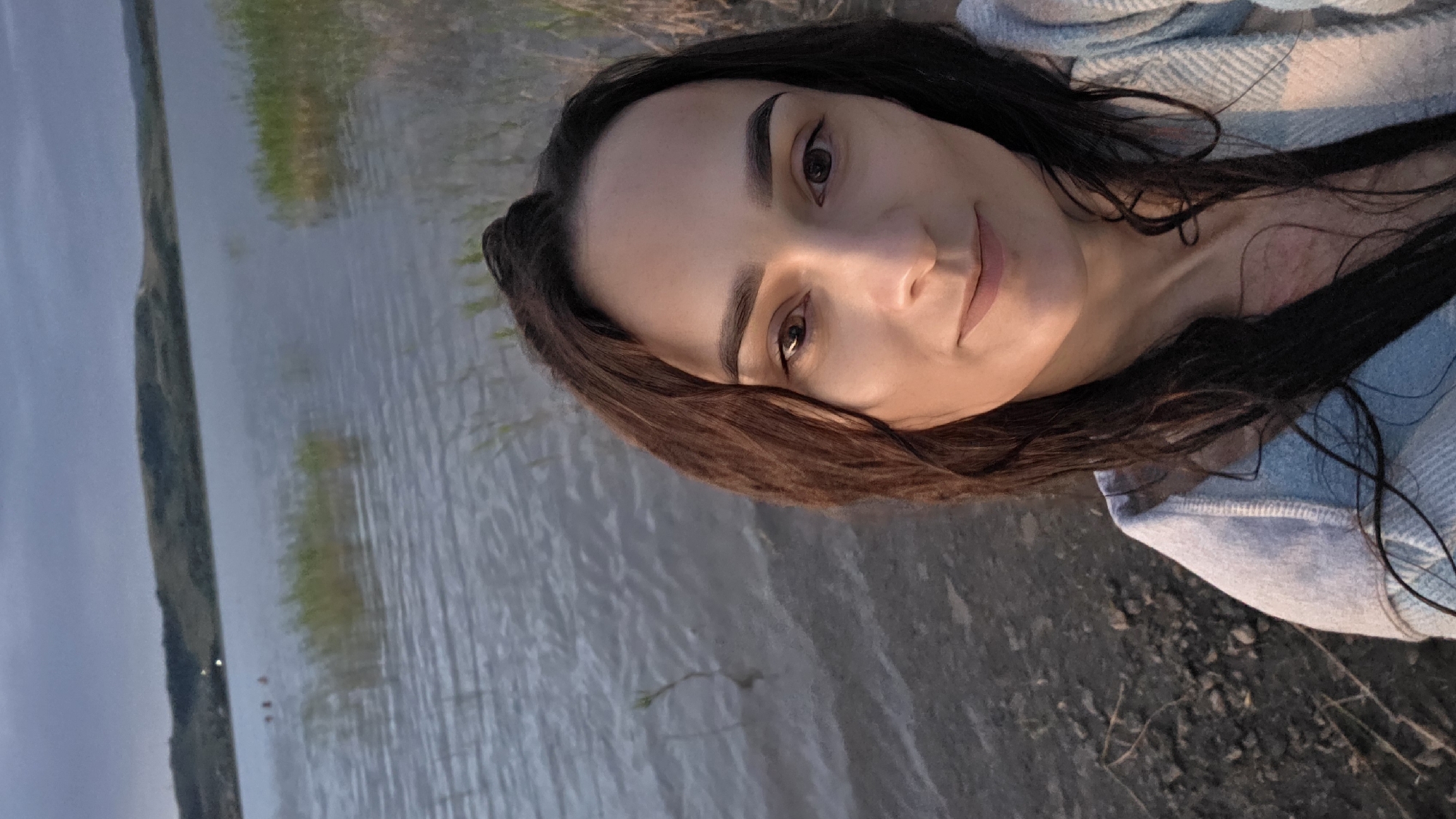 A selfie shows a young woman with dark brown hair and light brown eyes standing at the edge of a body of water. The water is choppy and a pale blue-grey, with reeds growing in patches. In the background, a low, rolling hill is visible under a cloudy sky. The ground in the foreground is rocky and dark. The woman is wearing a light blue, textured top. Her hair is wet and falling around her shoulders. The image appears to have been taken at dusk. The location is identified as Kupoyarovo, Russia.