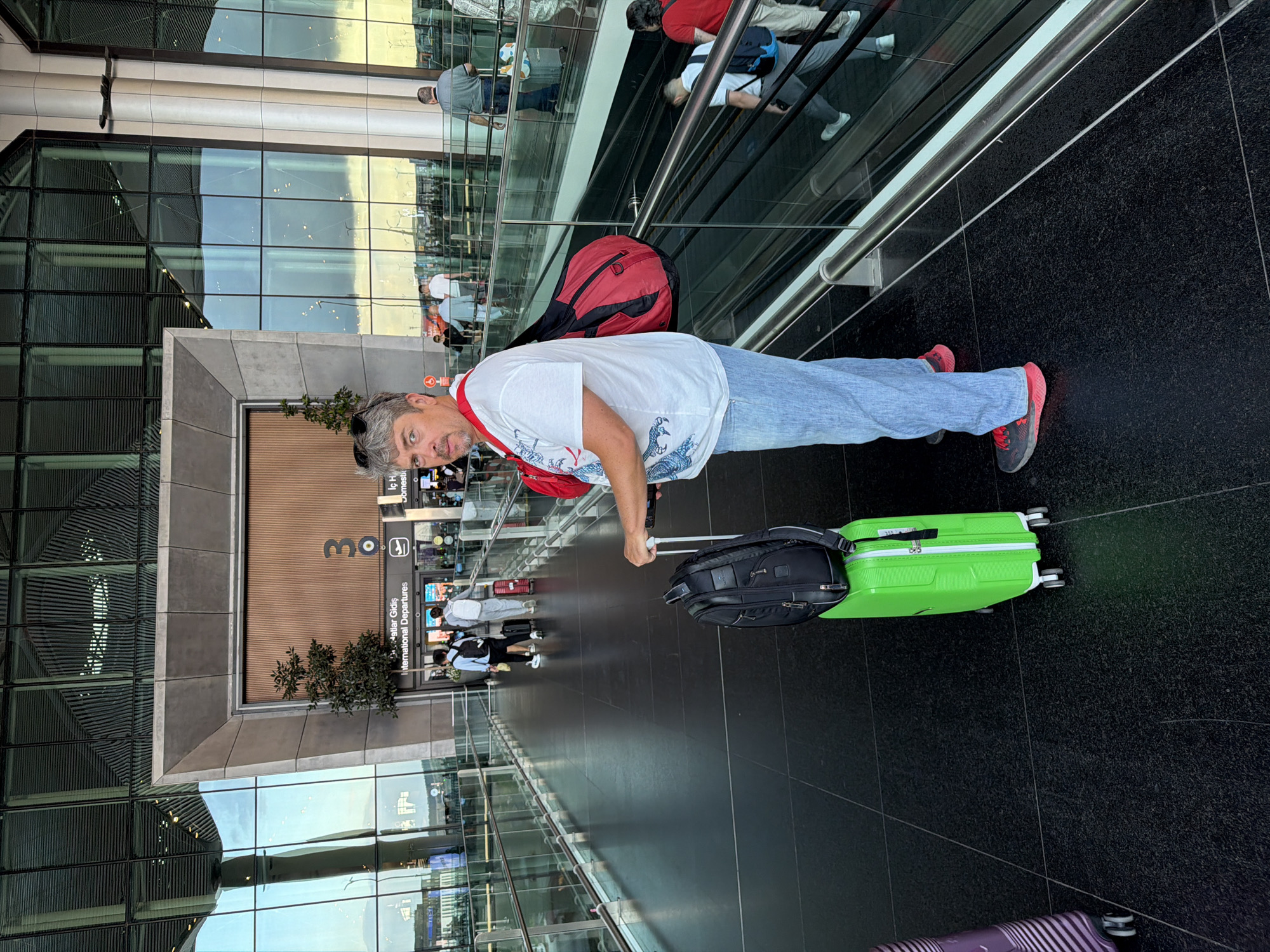 A man stands in an airport terminal, looking at the camera. He is wearing a white t-shirt with a blue dragon design, blue jeans, and red-soled sneakers. A red bag is slung over his right shoulder, and a black backpack sits on top of a bright green suitcase in front of him. The man is positioned in the foreground of the image, with an airport concourse stretching out behind him. Other travelers are visible in the background, some walking and others on an escalator. The terminal features large glass windows and modern architecture. This scene is located at Istanbul Airport, in Arnavutköy, Turkey, which is the primary international airport serving Istanbul.