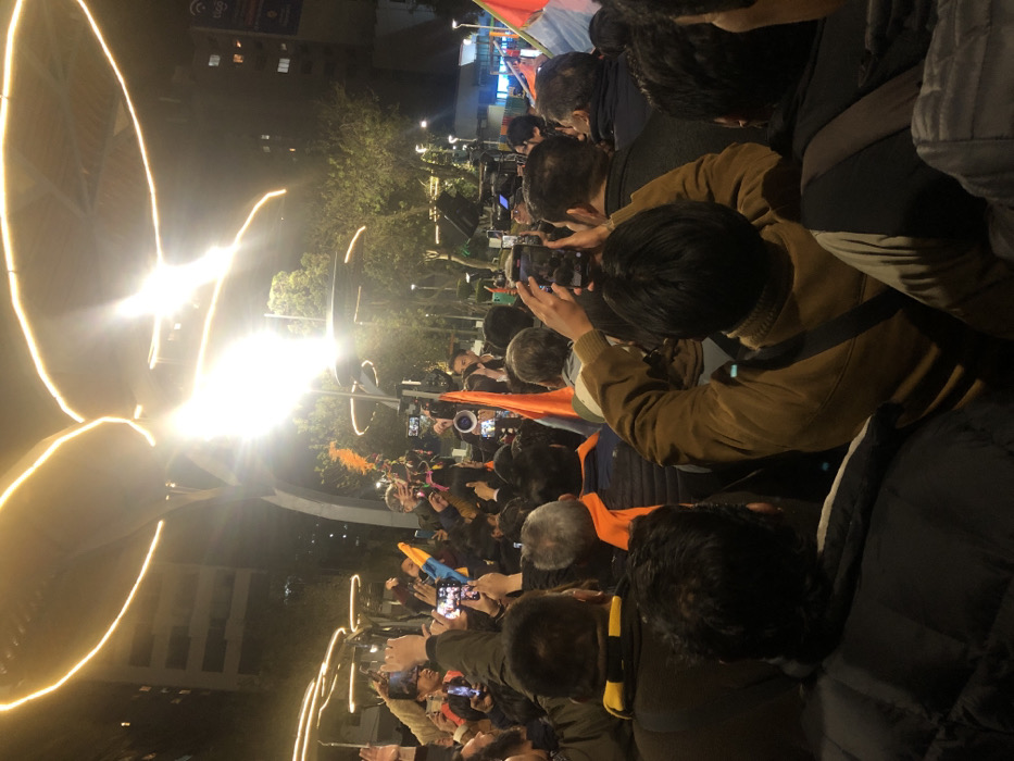 A crowd gathers at night, illuminated by bright, arcing lights. Many individuals are holding up their phones, seemingly capturing a scene unfolding before them. An orange flag and a megaphone are visible amidst the throng. Buildings and trees are faintly visible in the background, suggesting an urban setting. The focus is on the collective action and observation of the event.
