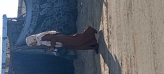 A female individual walks from left to right across an unpaved, dusty, and uneven dirt path in Khan Yunis, Palestinian Territory. She wears a light-colored hijab, dark sunglasses, and a long, dark brown dress or abaya. In her left hand, she holds a dark object, likely a mobile phone, and a small dark bag or item in her right hand. Her left foot is forward, with her right foot in motion. Behind her is a makeshift structure composed of dark tarpaulins or blankets, one of which displays a faint floral or patterned design. A lighter fabric or material is visible along the top edge of this structure. Strong natural sunlight illuminates the scene, casting a prominent shadow of the individual onto the ground to her left.