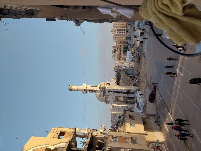 The image captures a wide-angle, elevated view of an urban landscape exhibiting extensive damage, likely from conflict or disaster. The photo is taken from what appears to be a damaged building, looking out over a street and surrounding structures under a clear blue sky, indicating daytime.
In the immediate foreground, at the top of the image (if oriented correctly, which is currently rotated 90 degrees counter-clockwise), a severely damaged concrete ceiling or overhang is visible, with exposed rebar and crumbling sections. A light yellow or beige fabric, possibly a towel or cloth, hangs down on the right side, partially obscuring the view. A dark, thick cable or wire curves into the frame from the right.
Below this foreground obstruction, the scene opens up to a wide street that runs from the left foreground towards the right background. The street appears paved and has faint markings, possibly for crosswalks or lane divisions. Numerous people are visible on the street, some walking individually or in small groups, while others are riding bicycles. Their clothing styles are varied, but many appear in dark attire.
To the left of the street (viewer's left, if oriented), the urban environment is characterized by widespread destruction. Buildings are heavily damaged or reduced to rubble, with skeletal remains of concrete and brick structures. One prominent building in the center-left is a mosque, easily identifiable by its distinct minaret and dome. The mosque also shows signs of damage, with visible pockmarks and holes on its walls and minaret, and parts of its structure appear to be crumbling. Large piles of debris and rubble are scattered between the damaged buildings and the street.
Further in the background, beyond the immediate devastation, more buildings can be seen, some appearing less damaged or even intact, suggesting a gradient of destruction across the area. The overall impression is one of a city grappling with the aftermath of significant damage, where daily life continues amidst widespread ruin. No discernible text is visible in the image.