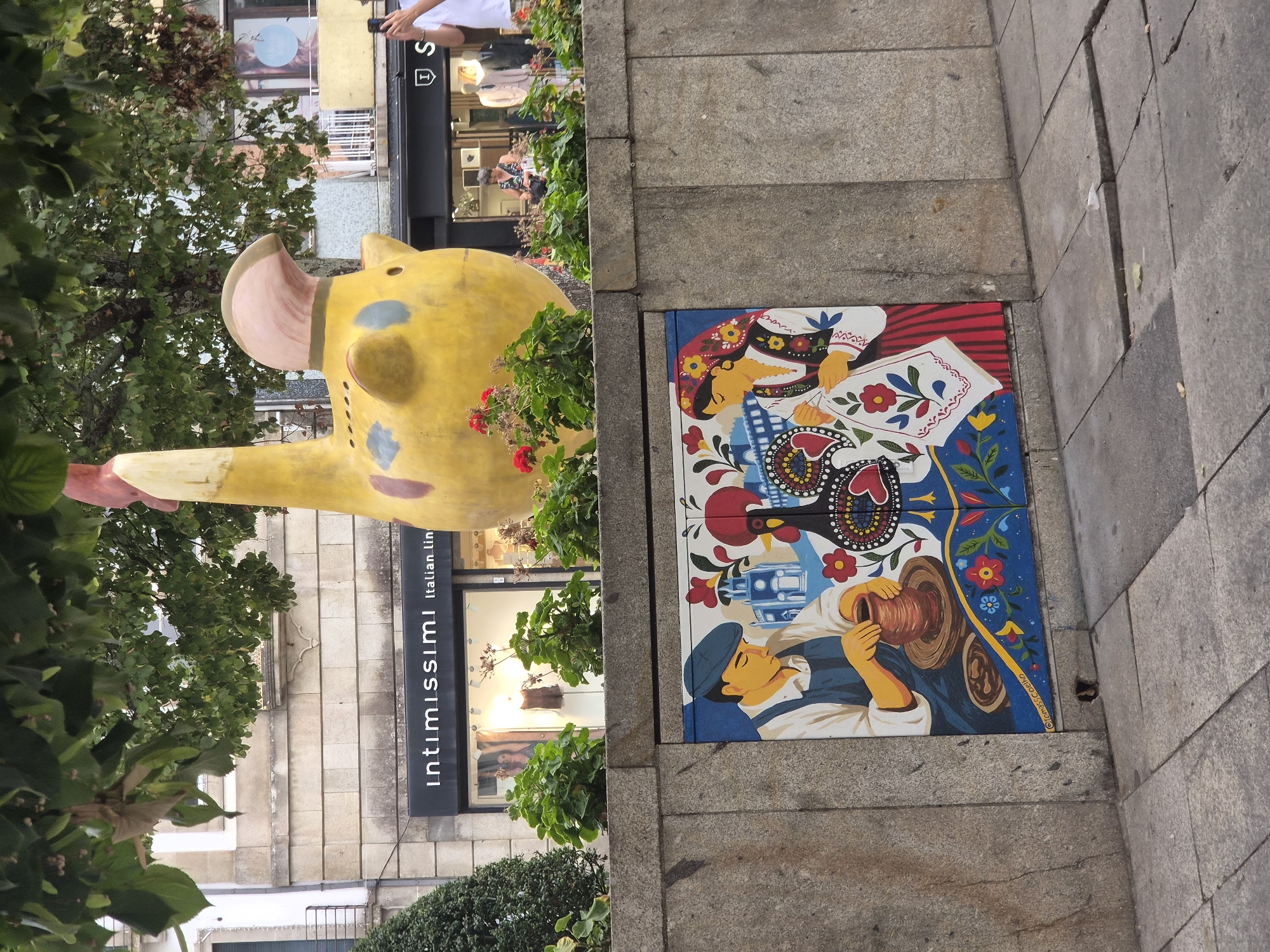 The image captures an outdoor scene in Barcelos, Portugal, featuring a large, yellow, decorative rooster sculpture prominently placed above a stone wall. The wall itself is adorned with a vibrant mural depicting traditional Portuguese folk art. The mural shows two figures: a man with a blue cap and apron, engaged in pottery making, and a woman in traditional attire with a headscarf, working with fabric. Between them, a stylized rooster is depicted, surrounded by colorful flowers and architectural elements. In the background, storefronts of businesses, including "Intimissimi Italian Lin," are visible, with mannequins displaying clothing. A person is partially visible on the right side of the frame, holding a smartphone. Green foliage from overhead trees partially obscures the top of the sculpture and buildings. The ground in the foreground consists of grey paving stones, some of which are cracked. The overall impression is a blend of public art, commercial activity, and a glimpse into local culture.