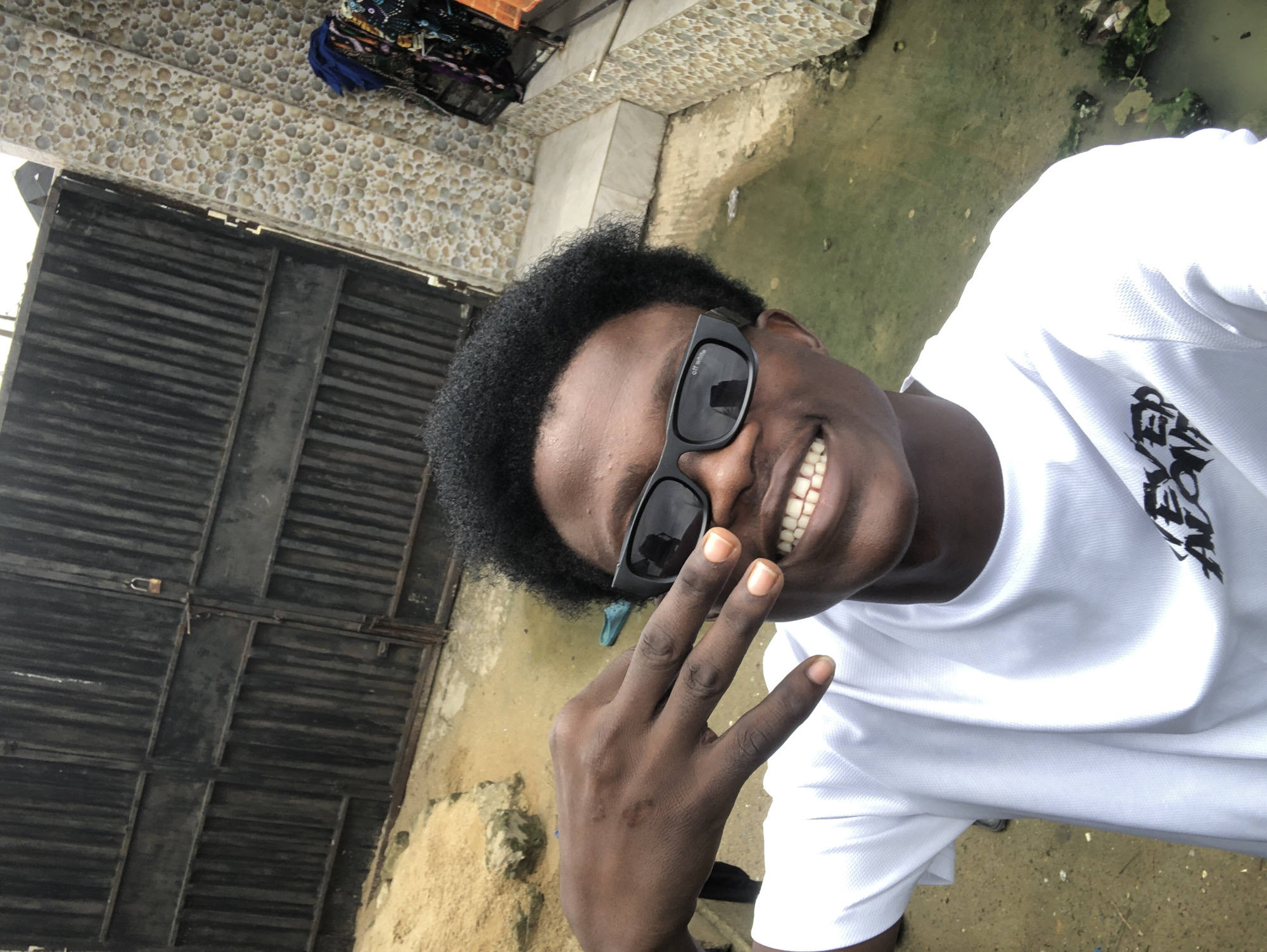 A young Black male, wearing a white t-shirt and black sunglasses, poses for a selfie in what appears to be an outdoor urban setting in Ado, Nigeria. He is flashing a peace sign with his left hand. Behind him, a large, black metal gate with a padlock is visible, along with a textured, light-colored wall. The ground is a mix of sand and what looks like standing water, suggesting a potentially unpaved area or a recent rain. The overall impression is a candid snapshot taken in a somewhat rustic or undeveloped part of a city.