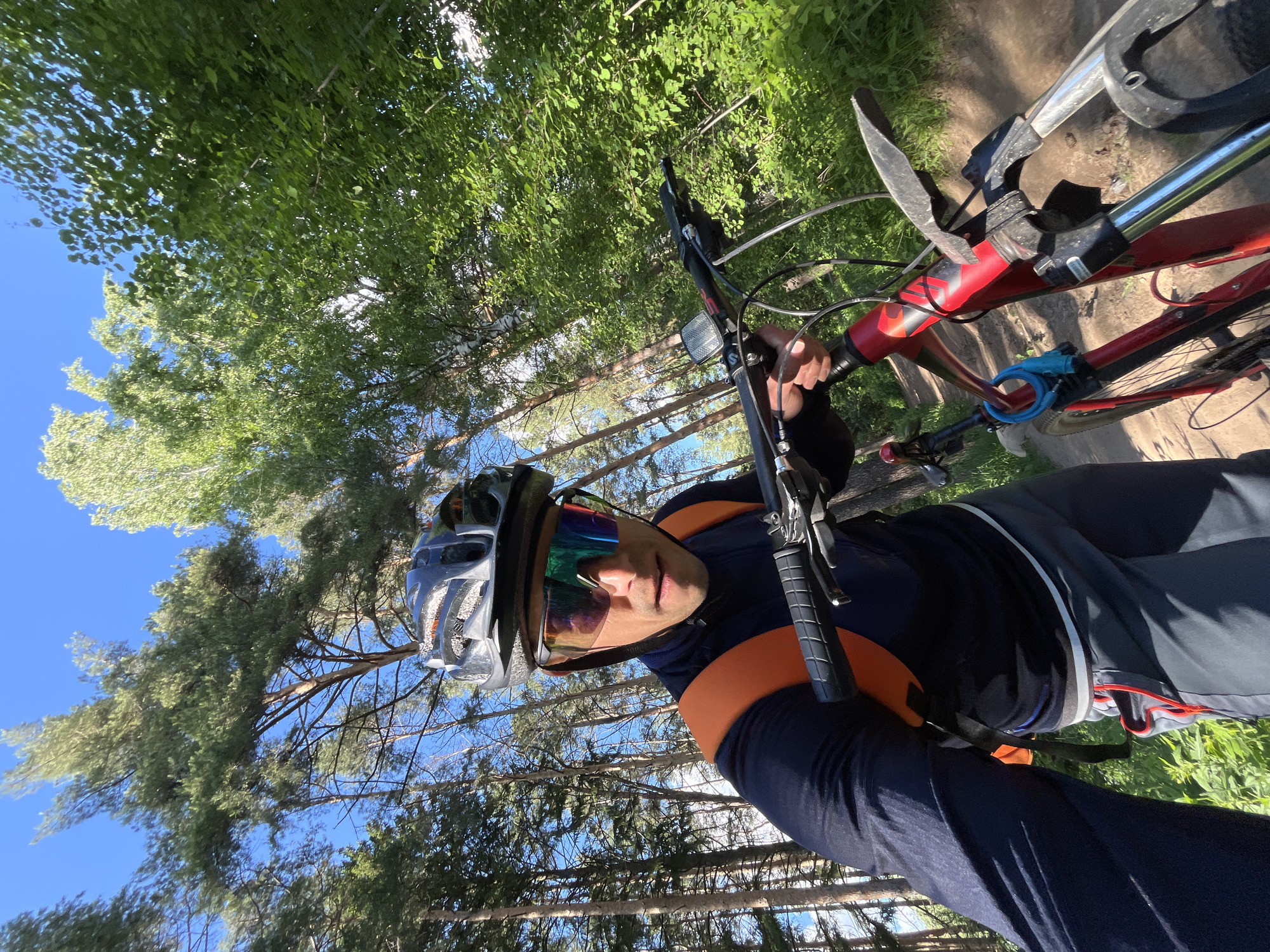 A man is standing outdoors with a red and silver mountain bike, appearing to have taken a selfie. He is wearing a helmet with a silver and gray pattern and reflective sunglasses. He has on a long-sleeved navy blue shirt with orange shoulder accents. The background consists of a forest with tall, thin trees and a bright blue sky visible between the branches. The image seems to have been taken in a wooded area in the city of Chaikovsky, Russia. The trees and ground suggest a natural, possibly park-like environment.