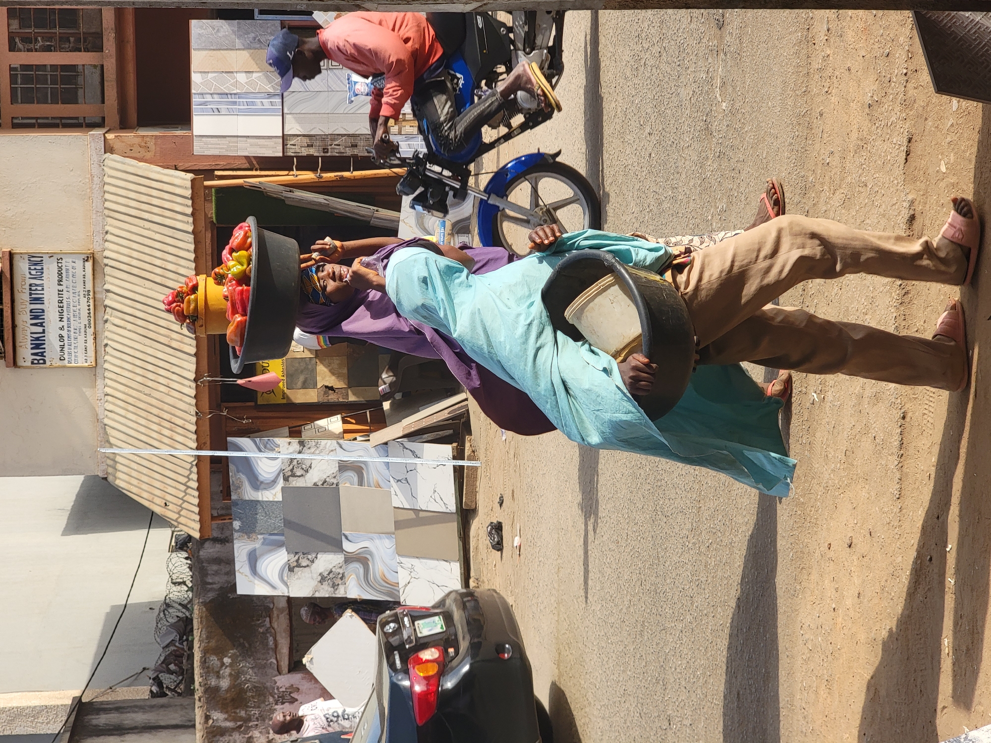 The image shows a woman walking on a street in Maiburiji, Nigeria, balancing a large bowl of colorful peppers on her head. She is wearing a light blue dress and tan pants with pink sandals. She has a smaller black bowl in her right hand. A man on a blue motorcycle is nearby, wearing a salmon-colored shirt and blue cap. To the side is a building with a sign that reads "BANKLAND INTER AGENCY," advertising Dunlop & Nigerite products. In front of the building are tiles, and there is a black car parked partially in the shot. The environment suggests an urban setting with buildings and a paved road.