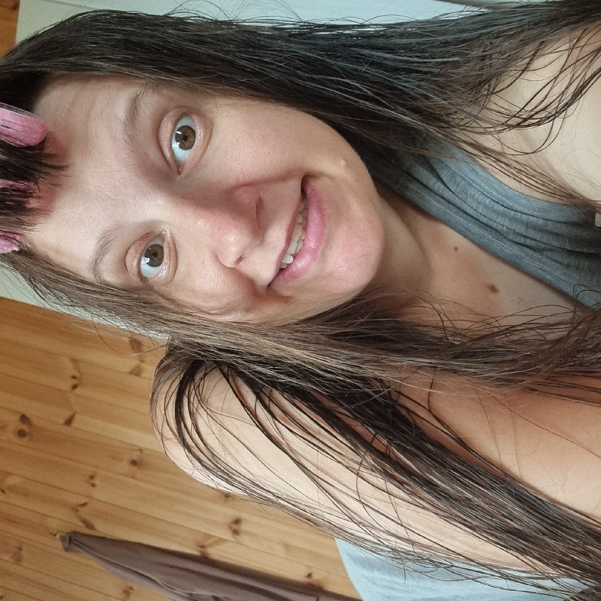 A woman with wet, dark brown hair styled with pink rollers is seen from the chest up, looking directly at the camera. She is wearing a gray tank top. Her right arm is bent and rests behind her head, with her hair draped over it. The background consists of light-colored wooden paneling, and a brown garment hangs on the left side of the frame. The scene is indoors, likely in a domestic setting in Dunnae-myeon, South Korea.