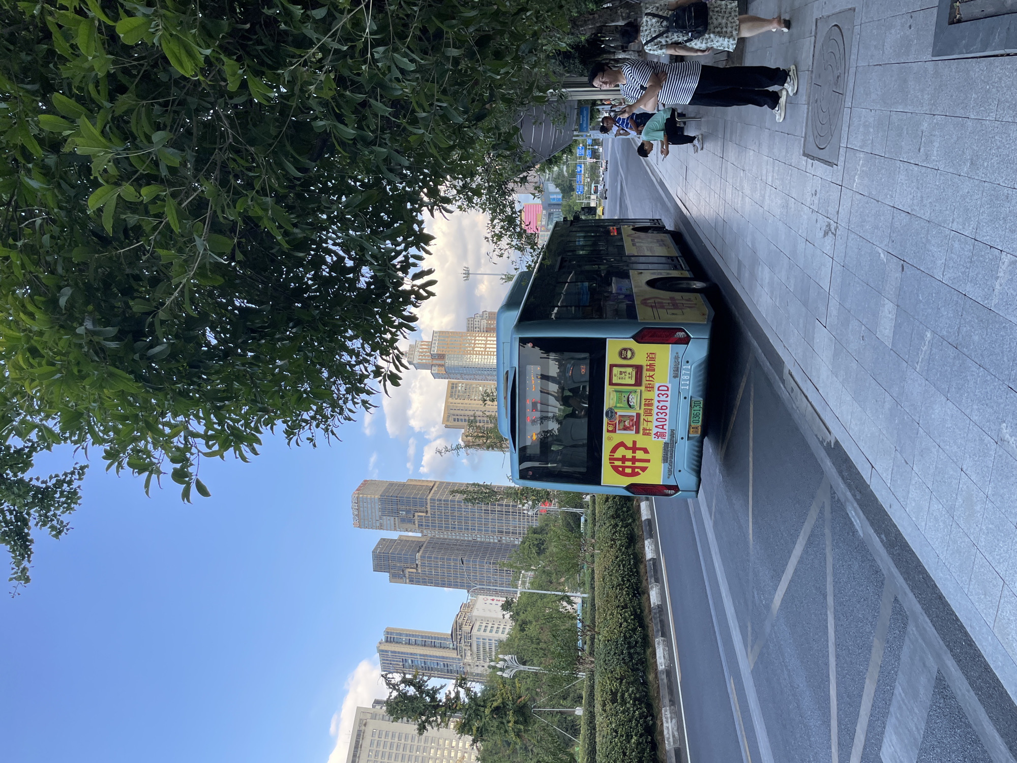 A light blue city bus is parked at a bus stop on a street in Nan'an District, China. The bus has advertisements on its rear, including Chinese characters and product images. Several people are standing and sitting on the sidewalk, waiting for the bus. The sidewalk is made of gray concrete tiles, and there is a green lawn and some trees separating the sidewalk from the street. In the background, there are modern skyscrapers and buildings, indicating an urban environment. The sky is blue with some scattered clouds. The scene appears to be during the daytime.