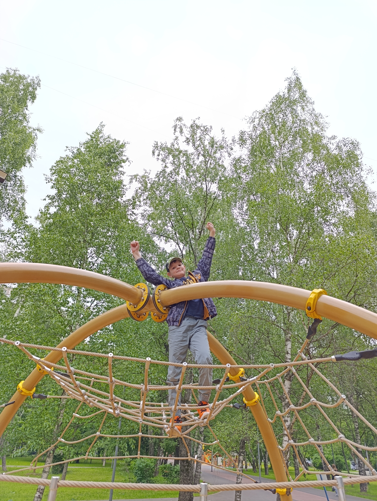 A young boy is captured in an outdoor playground setting, standing triumphantly on a rope climbing net. The boy, who appears to be of elementary school age, is dressed in a dark long-sleeved shirt with light patterns, grey shorts, a baseball cap, and distinctive orange and black sneakers. He has both arms raised above his head, fists clenched, in a gesture of excitement or accomplishment.
The climbing structure is robust, featuring large, curved, yellow or tan metal tubes that form the main framework, with the intricate rope netting stretched between them. Yellow clamps and black connectors secure various parts of the structure.
The background is dominated by lush green foliage from numerous birch trees, indicating a park or heavily treed area. The sky above is bright and uniformly white, suggesting an overcast daytime. Portions of paved pathways, grass, and other distant playground equipment are visible around the base of the structure. The low camera angle emphasizes the boy's position atop the net, creating a dynamic sense of achievement within a vibrant, natural environment, consistent with a park in Moscow, Russia. No discernible text is visible on the boy's clothing or within the scene.