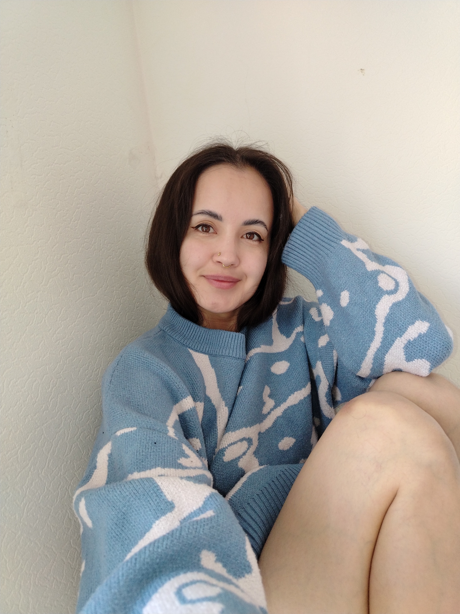 A close-up, chest-up portrait features a young woman with shoulder-length dark brown hair and warm brown eyes, looking directly at the viewer with a gentle, engaging smile. She has delicate eyeliner, a small gold hoop nose ring on her left nostril, and a small gold stud on her right. She is wearing a cozy-looking, light blue knitted sweater with an abstract white pattern. Her right arm is bent with her hand resting on the back of her head or neck, while her left arm is bent and resting lower. Her bare legs, with knees drawn up, are visible in the lower right portion of the frame. The woman appears to be seated indoors, possibly on the floor or a low surface. The background is a plain, light off-white or beige wall with a subtle textured pattern. The lighting is bright and even, suggesting daytime. No text is visible in the image. The overall mood is calm and friendly. This indoor scene is contextualized as being in Noviy, Russia.
