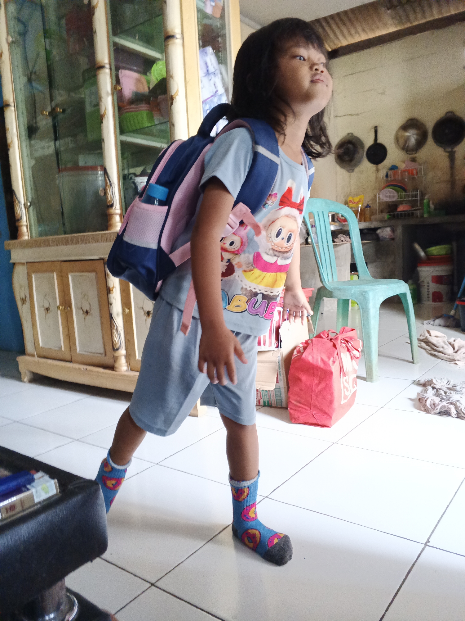 A young Indonesian girl with dark hair stands on a tiled floor in what appears to be a home in Warungkiara, Indonesia. She is wearing a blue t-shirt with a cartoon character, gray shorts, and colorful socks with donuts. A blue and pink backpack is on her back, and she is looking upwards and to the right with her mouth slightly open. In the background, there is a large wooden cabinet with glass doors, a blue plastic chair, a red bag, and shelves with various items. The environment suggests a cluttered but lived-in space.