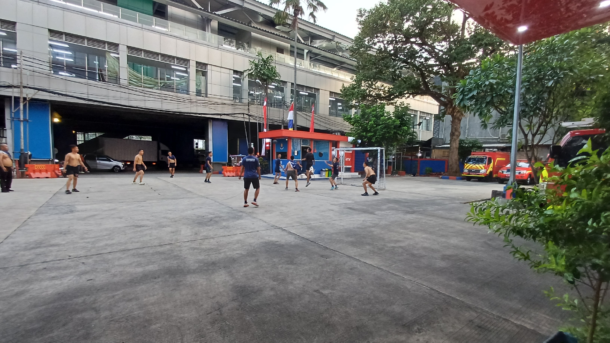 A group of men are playing soccer on a concrete court in front of a multi-story building. The building has a modern facade with large windows and a green roof. In the background, a red fire truck and another red vehicle are parked. The court is marked with lines, and there is a soccer goal at one end. Several men are in mid-action, one is jumping to head the ball, and others are running or standing. The scene appears to be in a semi-urban or industrial area. The specific location in the city of South Jakarta, Indonesia, is indicated by the presence of these vehicles and the architecture of the building.