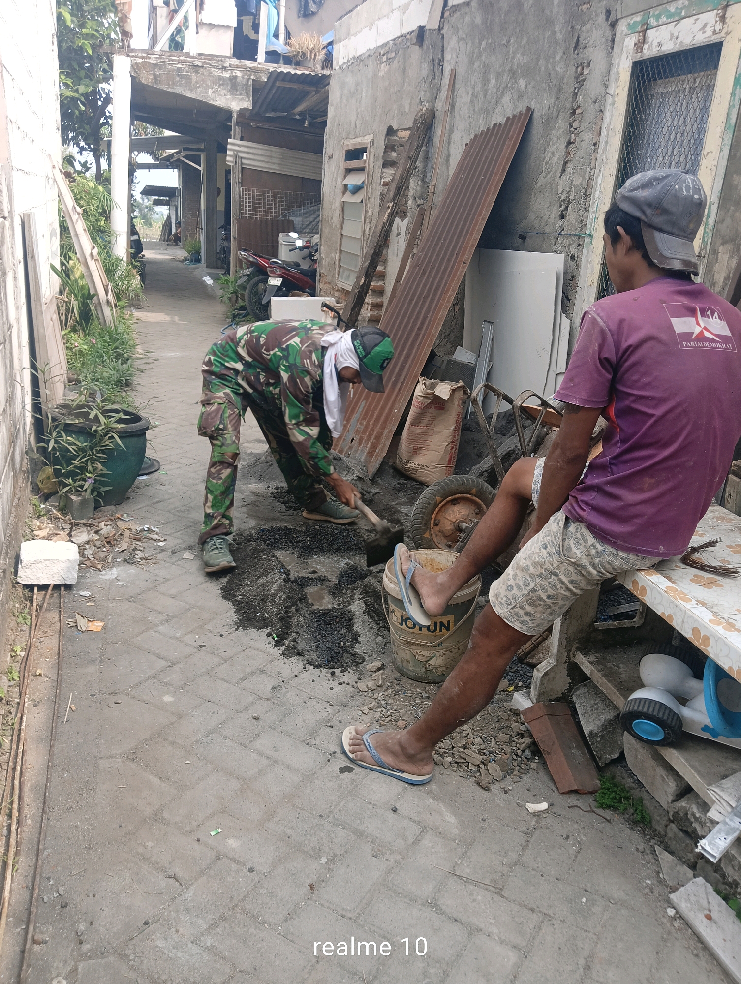 In a narrow alleyway in Batu, Indonesia, two men are present. One man, wearing camouflage pants and a green and white headscarf, is crouched down, using a shovel to work with a pile of dark material on the paved ground. The other man, wearing a purple t-shirt and patterned shorts, sits on a white table to the right of the scene. He is looking away from the camera. In the background, buildings with corrugated metal roofs and unfinished concrete walls are visible, along with some potted plants and parked motorcycles. A wheelbarrow and several buckets are also scattered around the alley. The overall impression is one of a work in progress in a somewhat cluttered urban environment.