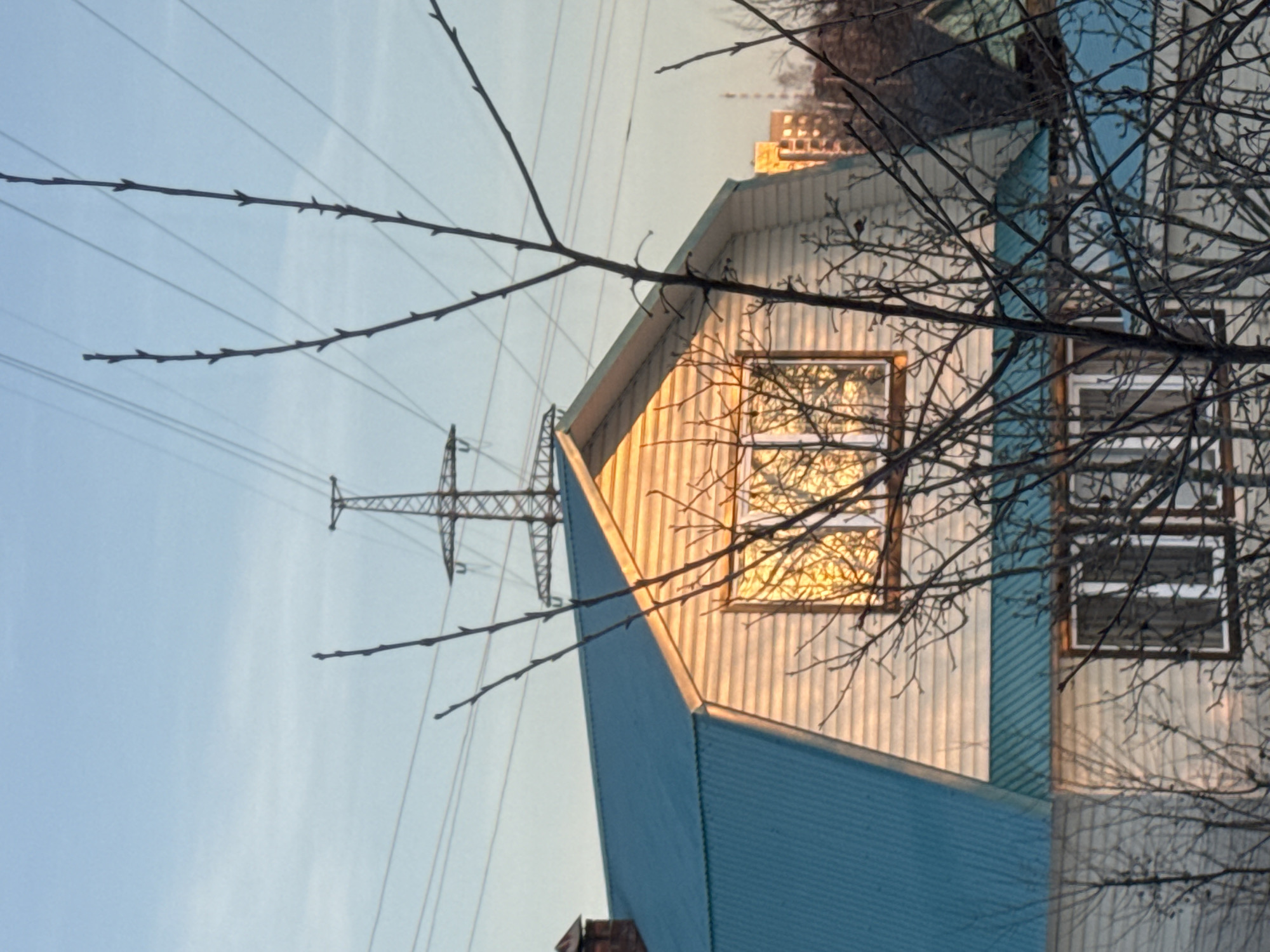 The image shows the side of a house in Chaikovsky, Russia. The house has a blue and white exterior with windows reflecting sunlight. A power line tower is visible in the background, partially obscured by tree branches. The sky is light blue.