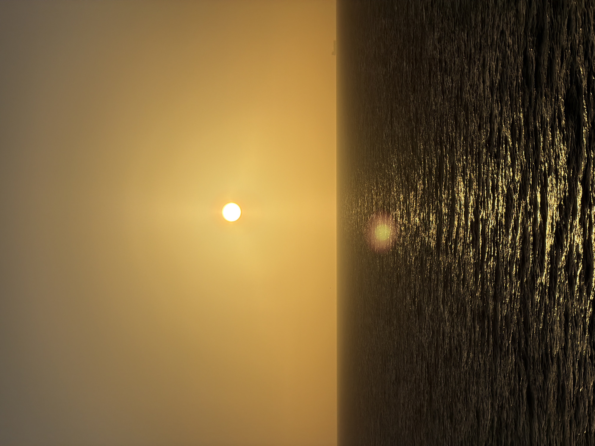 The image presents a scene with the sun positioned on the left side against a light orange background. To the right, there is a dark, textured surface with light reflecting off it. The scene is divided by a vertical line. It is hard to tell where in Unknown, Unknown this is as there is nothing to identify this location.