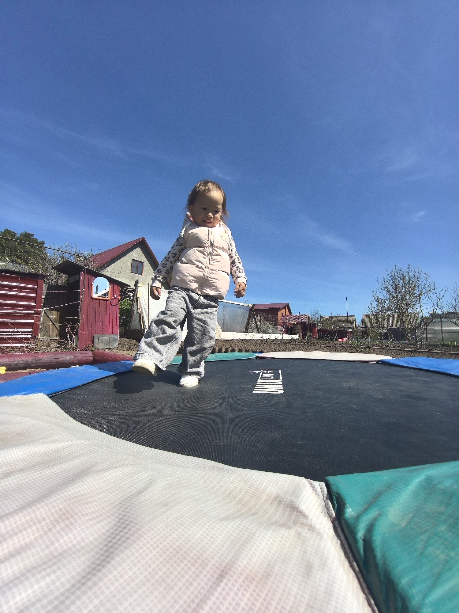 A toddler is standing on a trampoline in Noviy, Russia. The child wears a light-colored, patterned jacket, grey pants, and white shoes. The trampoline is round and has a safety pad with a white edge, blue trim, and a green border. The background shows several houses and sheds with red and brown siding. The sky is clear and blue with light clouds.