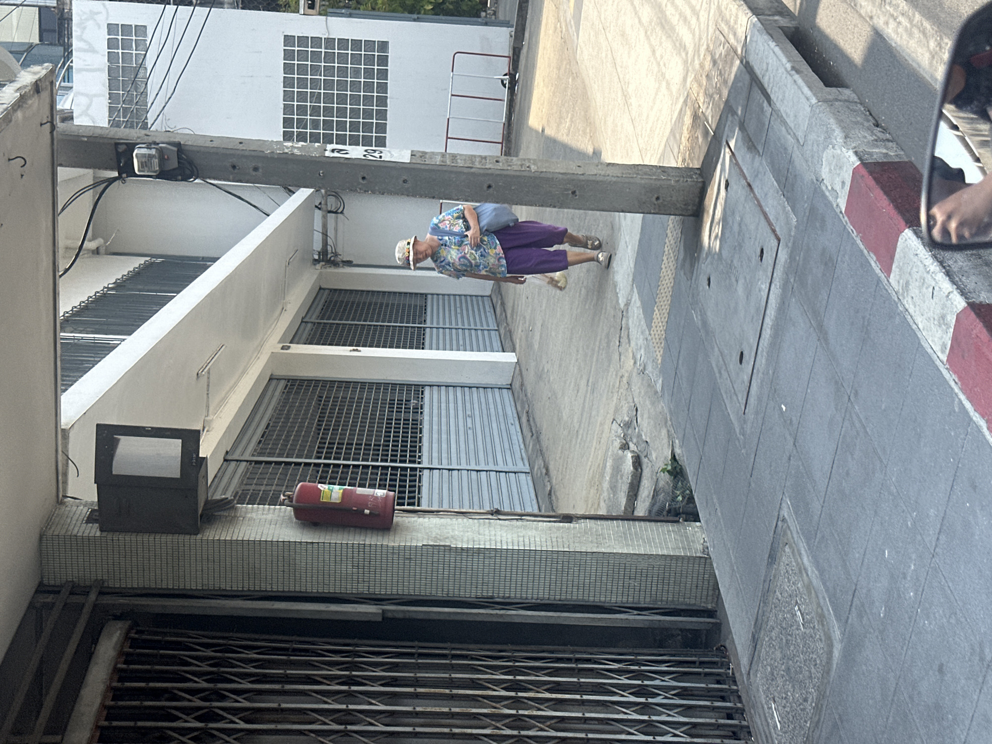 The image shows a street scene in Bangkok, Thailand. An elderly woman wearing a patterned shirt, purple pants, and a hat walks on the sidewalk. She is carrying a blue bag. To the left, a building has a metal security gate, a gray box, and a red fire extinguisher mounted on the wall above the gate. A concrete utility pole runs across the top of the frame. The sidewalk is made of gray tiles. A red and white curb borders the sidewalk on the right. The scene is generally urban, with the architecture typical of the city.
