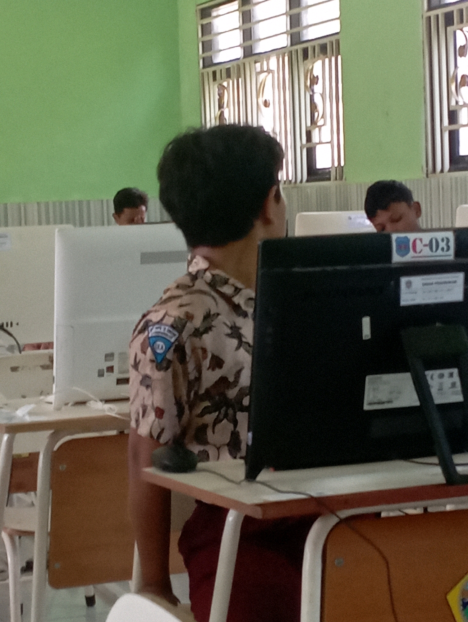 In a classroom setting in Banyuurip, Indonesia, two young students are visible, each seated at a computer desk. The student in the foreground, viewed from the back and side, wears a patterned school uniform shirt with a blue emblem on the left shoulder and dark red pants. This student is focused on a black computer monitor. To the right of this monitor, a white label displays "C-03" in red and blue lettering. Behind the foreground student, another student is partially visible, looking down at their own computer. The classroom environment includes white computer monitors, desks with brown surfaces and white metal frames, and a pale green wall. Windows with white bars are present in the background.