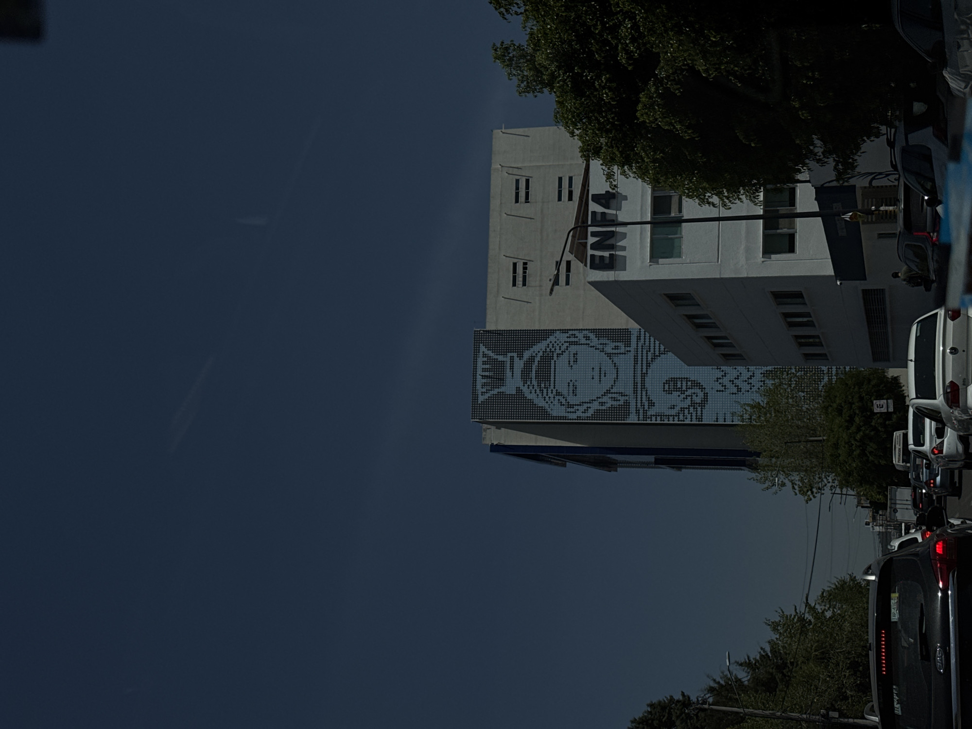 This image depicts an urban street scene in Puebla, Mexico, under a clear, deep blue sky, possibly in the late afternoon or approaching dusk. On the left side of the frame, a multi-story white building prominently displays the word "ENFA" written vertically in dark letters on its facade. Below this building, another structure features a large, pixelated or mosaic-style mural of a face, which appears to be that of a woman or a stylized figure, rendered in light tones against a darker grid. Additional buildings are visible in the background on the left.
To the right, a street is occupied by a line of vehicles, suggesting traffic. A white van or SUV is seen in the mid-ground, followed by a black car with visible taillights in the foreground. Trees with dense green foliage are scattered throughout the scene, bordering the street and overhanging some of the buildings, providing natural elements to the urban landscape. The focus is on the architectural features and the flow of city life, with no specific human activity other than driving discernible. The lighting creates distinct shadows and highlights, contributing to the atmospheric quality of the scene.