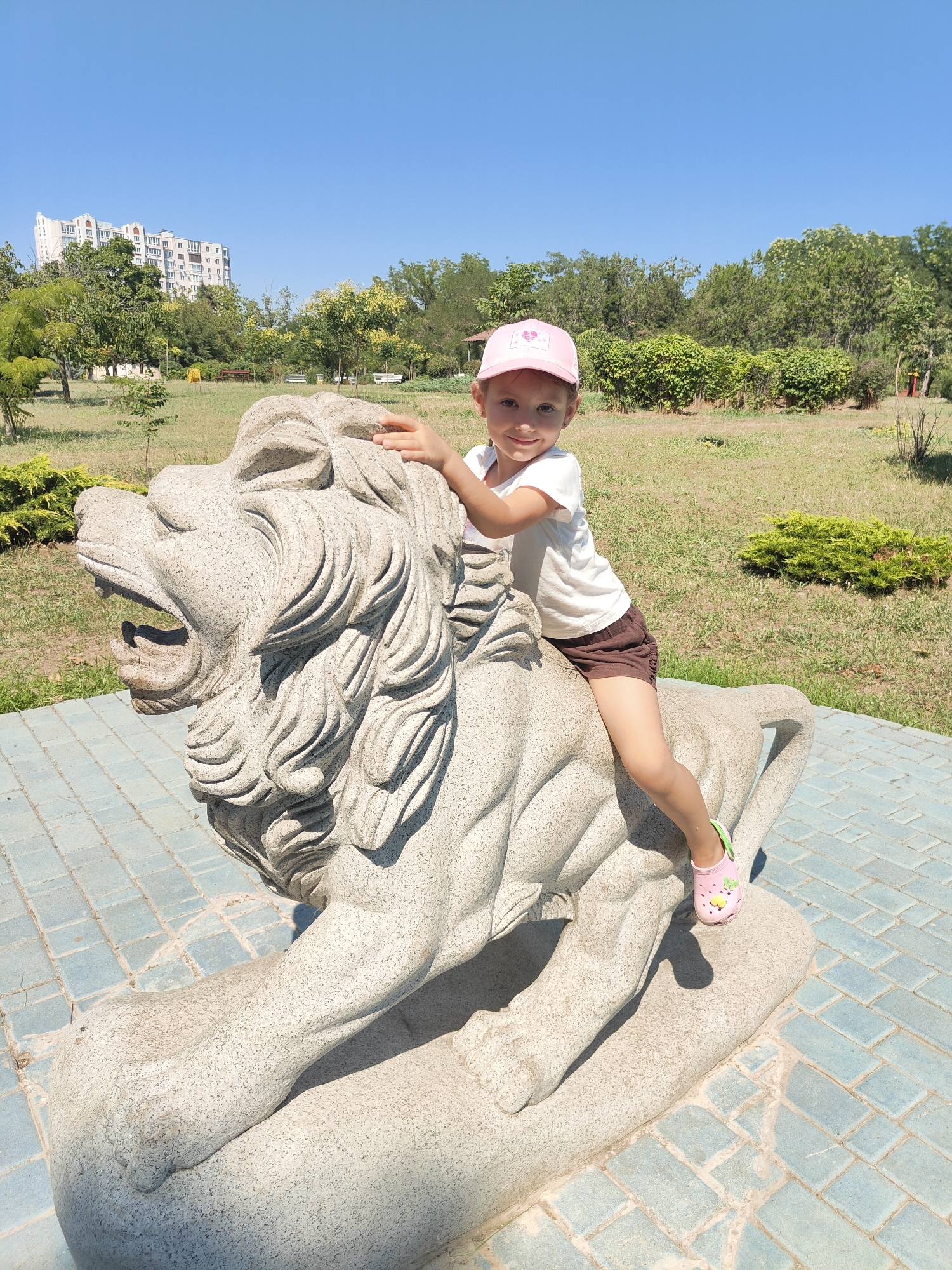 A young girl wearing a pink baseball cap, a white t-shirt, and brown shorts sits atop a large stone lion sculpture in a park. She is smiling and looking at the camera, with her left hand resting on the lion's mane. The lion sculpture is positioned on a blue tiled surface. In the background, there are trees, grass, and a tall, light-colored building with many windows. The sky is clear and blue.