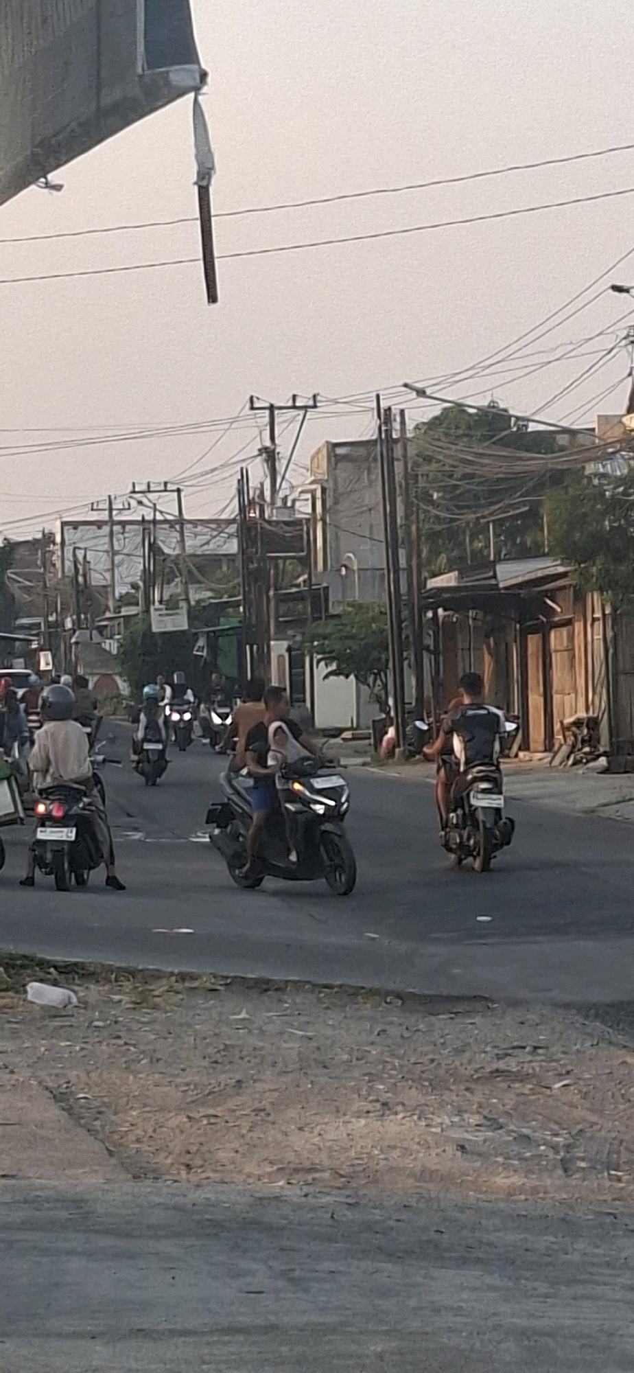 In the city of Desa Dibal, Indonesia, an overcast sky looms over a street scene populated by numerous motorcycles. The foreground is dominated by a paved road with a gravelly shoulder on the right. On the left side of the road, a concrete structure with a rough, textured surface is visible, from which a dark, elongated object hangs by a white strap, suspended over power lines.
Multiple motorcycles, some with riders and some without, are scattered across the road. A person on a motorcycle in the middle-left of the frame wears a light-colored shirt and a helmet. To their right, another motorcycle carries two individuals, one wearing a dark t-shirt and the other a light-colored garment. Further to the right, a motorcycle is ridden by a person in a dark t-shirt with white detailing on the back. The background reveals a row of buildings and densely packed power lines supported by utility poles. The overall impression is one of a busy street in a developed, possibly urban or suburban, area of Indonesia.