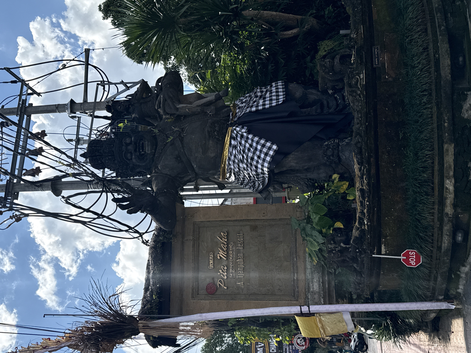 The image depicts a large, dark stone statue of a mythical figure in Ubud District, Indonesia. The statue is a menacing depiction with a large head, open mouth, and multiple arms, one of which appears to be holding a smaller figure. It is adorned with a gold sash and a black and white checkered cloth around its waist. The statue stands on a tiered, moss-covered base. To the left of the statue is a stone monument with the words "Pita Maha" and "A Tjampuhan Resort" inscribed on it, suggesting this is the entrance to a resort. A tall white pole with decorative elements stands in front of the monument. The scene is further characterized by lush green foliage surrounding the statue and monument. Overhead, a complex network of electrical wires and poles is visible against a bright blue sky with scattered clouds. A red octagonal stop sign hangs upside down near the base of the statue. The environment suggests a tropical urban setting, likely on a street or road, as a motorcycle is partially visible in the background on the left. The overall impression is one of local culture and spirituality integrated with modern infrastructure.