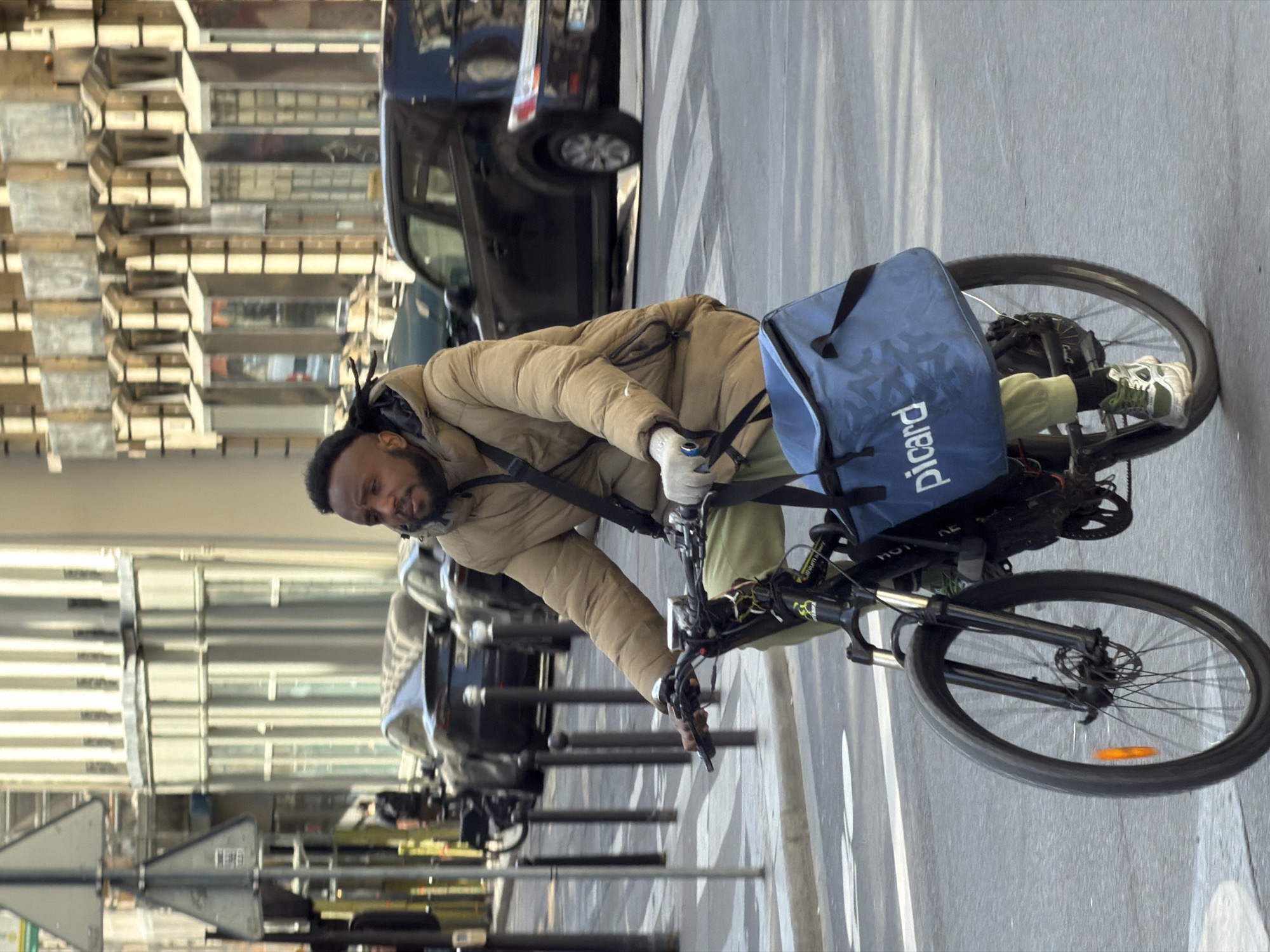 An upright, sunlit urban street scene captures a man riding an electric bicycle. The man, with a beard and dark dreadlocks/braids, is wearing a tan puffer jacket, light-colored gloves, and light green/yellow pants. He appears focused, leaning forward over the handlebars of his black electric bicycle, which features front suspension and disc brakes.
Attached to the front of the bicycle is a prominent blue delivery bag with the word "Picard" printed in white text, indicating he is likely a delivery driver.
The background on the left (viewer's left when upright) shows a multi-story building with numerous horizontal and vertical architectural elements, possibly balconies or windows, reflecting bright sunlight. On the right, a paved street or sidewalk is visible, with several dark-colored cars parked alongside, and a row of metal bollards or poles separating the road from what appears to be a pedestrian area or another part of the street. White lines are painted on the road surface.
The image was captured on a clear, sunny day, likely during the daytime, given the strong illumination and shadows. The presence of the "Picard" brand (a French frozen food retailer) and the urban setting strongly suggest the location is Paris, France.