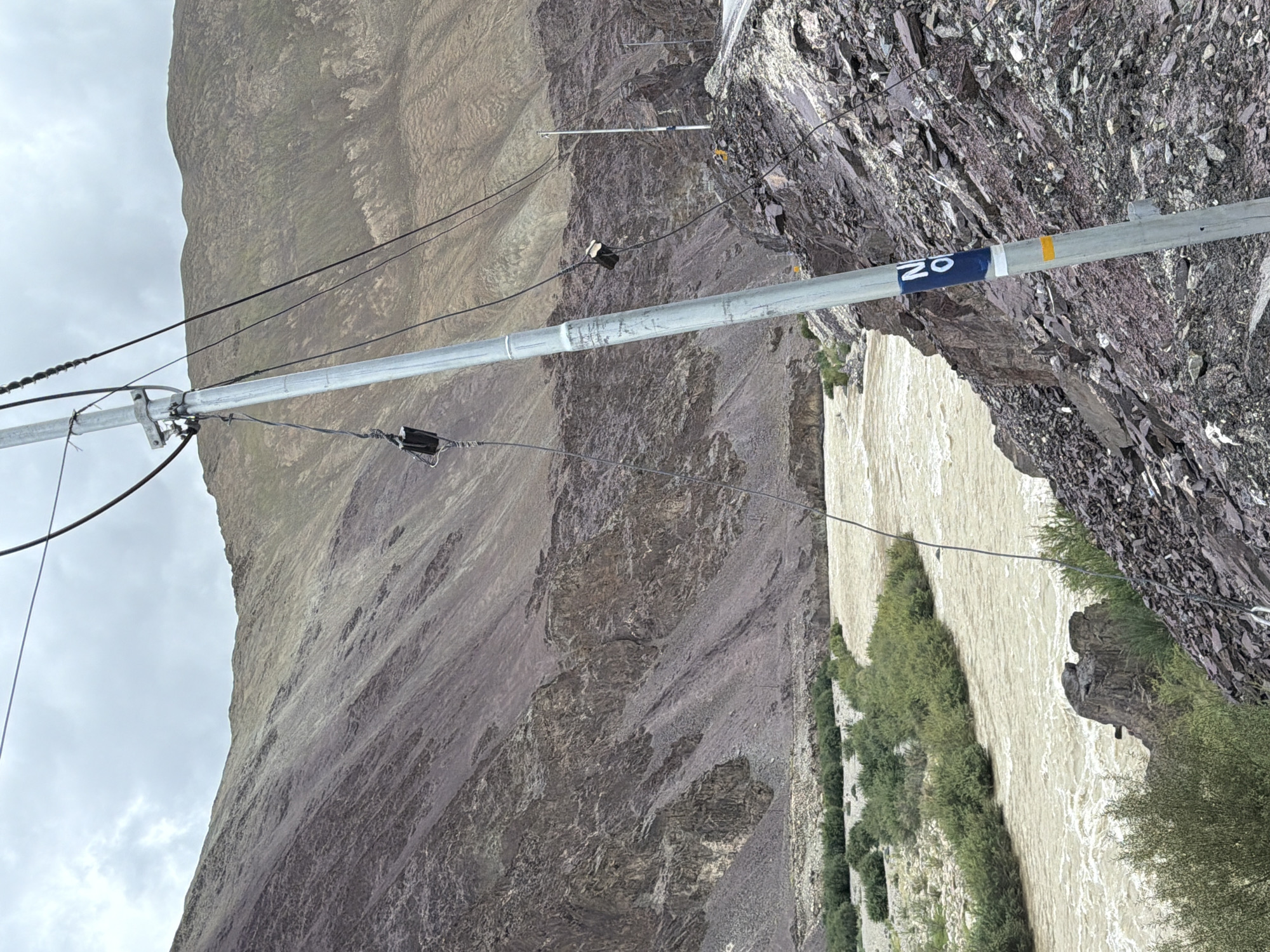 The image depicts a cloudy day in a mountainous region. In the foreground, a silver utility pole with numerous black cables stretches upwards and to the left, partially obscuring the view. The pole has a blue and white circular marker with the letters "NO" visible, and a yellow horizontal stripe below it. The pole is situated on a rocky, sloped terrain.
To the left of the pole, a wide, muddy river flows through a valley. The riverbanks are lined with green vegetation, including small trees and bushes. The valley is flanked by steep, rocky mountains with patches of green vegetation on their slopes. The mountains appear arid and weathered, with layers of sediment and rock formations visible.
In the background, more mountains rise, their tops shrouded in clouds. The sky is overcast with diffused light. A road is visible winding along the mountainside to the right of the utility pole, with another smaller pole visible in the distance. No people or animals are present in the scene. The location is identified as Kiari, India, suggesting a remote, natural landscape with infrastructure for utilities and transportation.