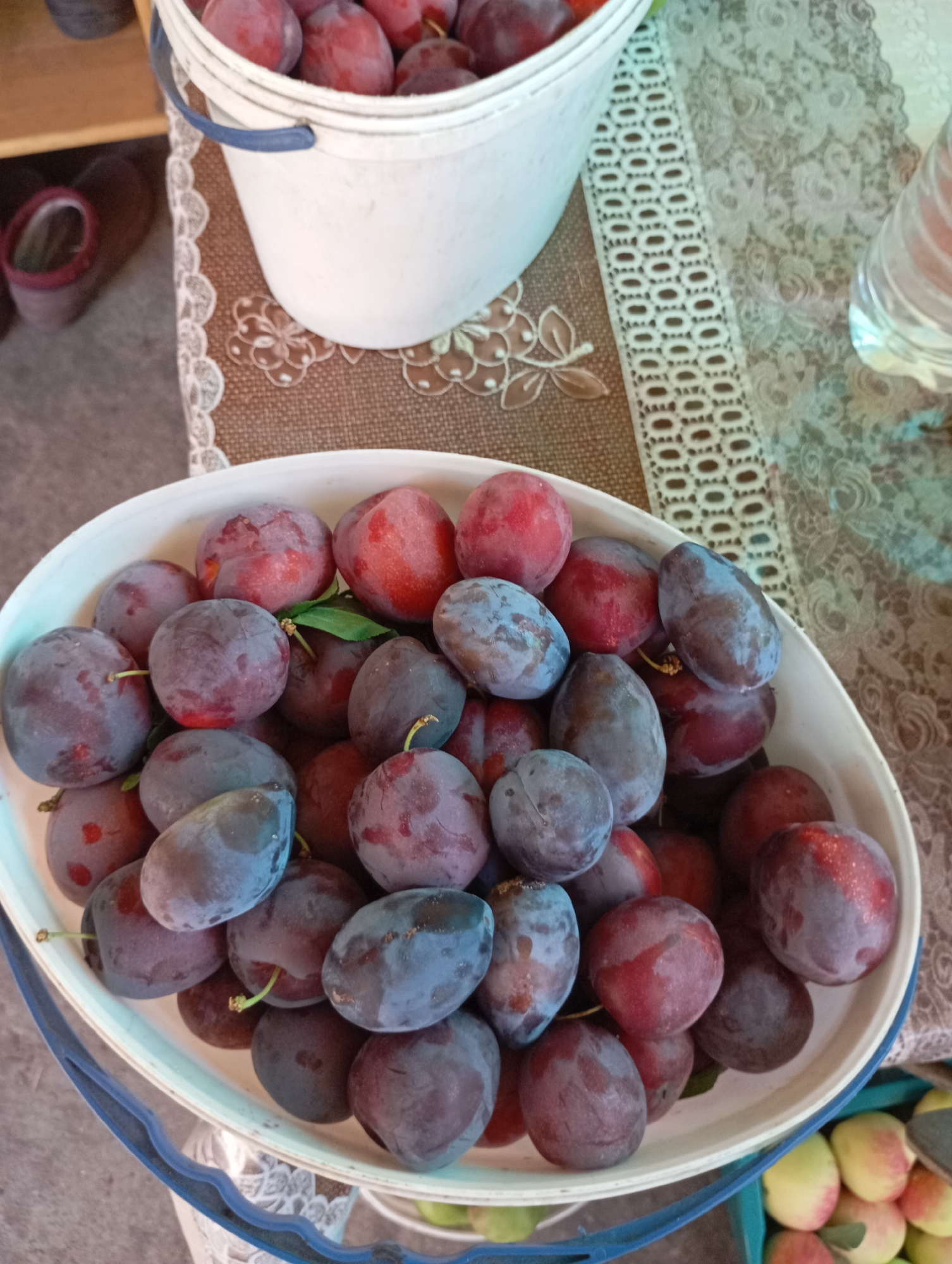 A high-angle shot shows a white bowl filled with ripe plums. The plums are a mix of deep purple and red, with some showing a dusty, bluish bloom. They are oval and round in shape, some with visible stems and a single green leaf. The bowl is white and round with a blue handle visible on the left side. The bowl sits on a table covered with a patterned cloth, featuring a brown base with a white lace trim. To the left of the bowl, a white bucket, also filled with plums, is partially visible. In the background, a clear glass bottle containing a liquid is on the right, and a pair of dark-colored shoes rests on the floor to the left. The overall scene suggests a harvest or preparation of fruit, possibly in an outdoor or semi-outdoor setting. The image was taken in Horishni Plavni, Ukraine.