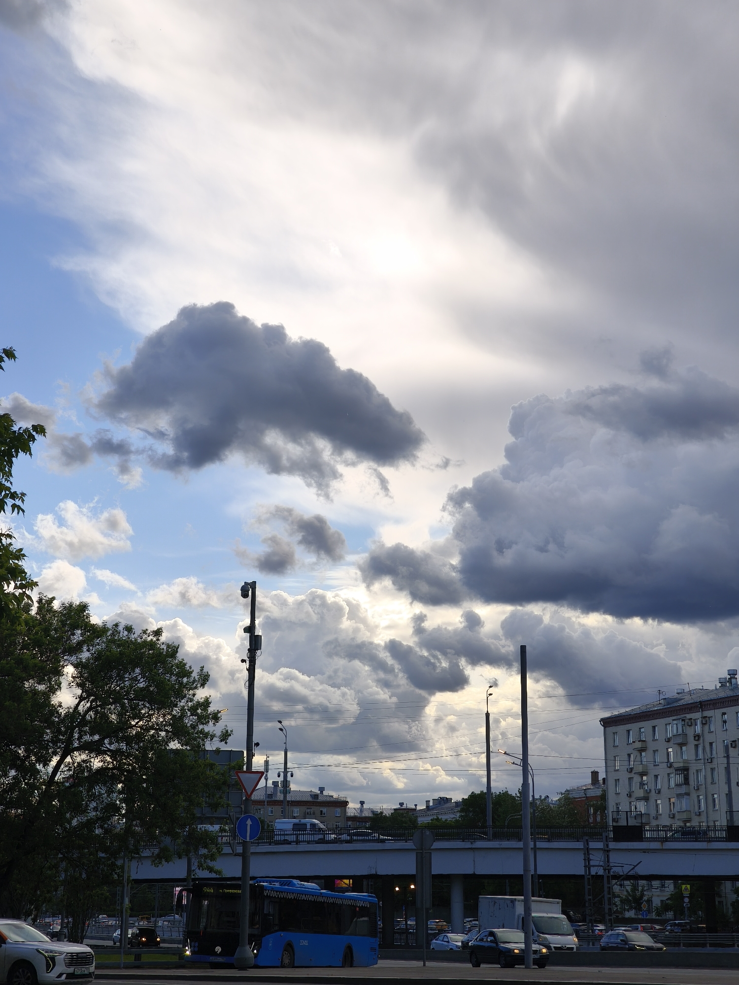 A street scene in Moscow, Russia on an overcast day. A blue bus and other vehicles are on the road. An elevated bridge carries traffic above. Buildings are in the background. Trees line the street. Power lines run across the sky. Cumulus clouds cover most of the sky.