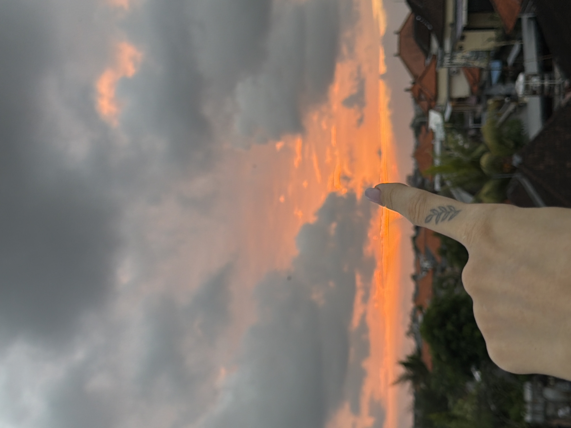 A close-up shot of a person's hand with a small leaf tattoo on their index finger is positioned in the foreground, pointing upwards towards a cloudy sky. The sky is dominated by dramatic sunset colors, with vibrant oranges and pinks peeking through dark gray clouds. Below the sky, a blurred cityscape of North Kuta, Indonesia, is visible, featuring terracotta-tiled rooftops and lush green foliage. The overall mood is serene and highlights the beauty of a tropical sunset.
