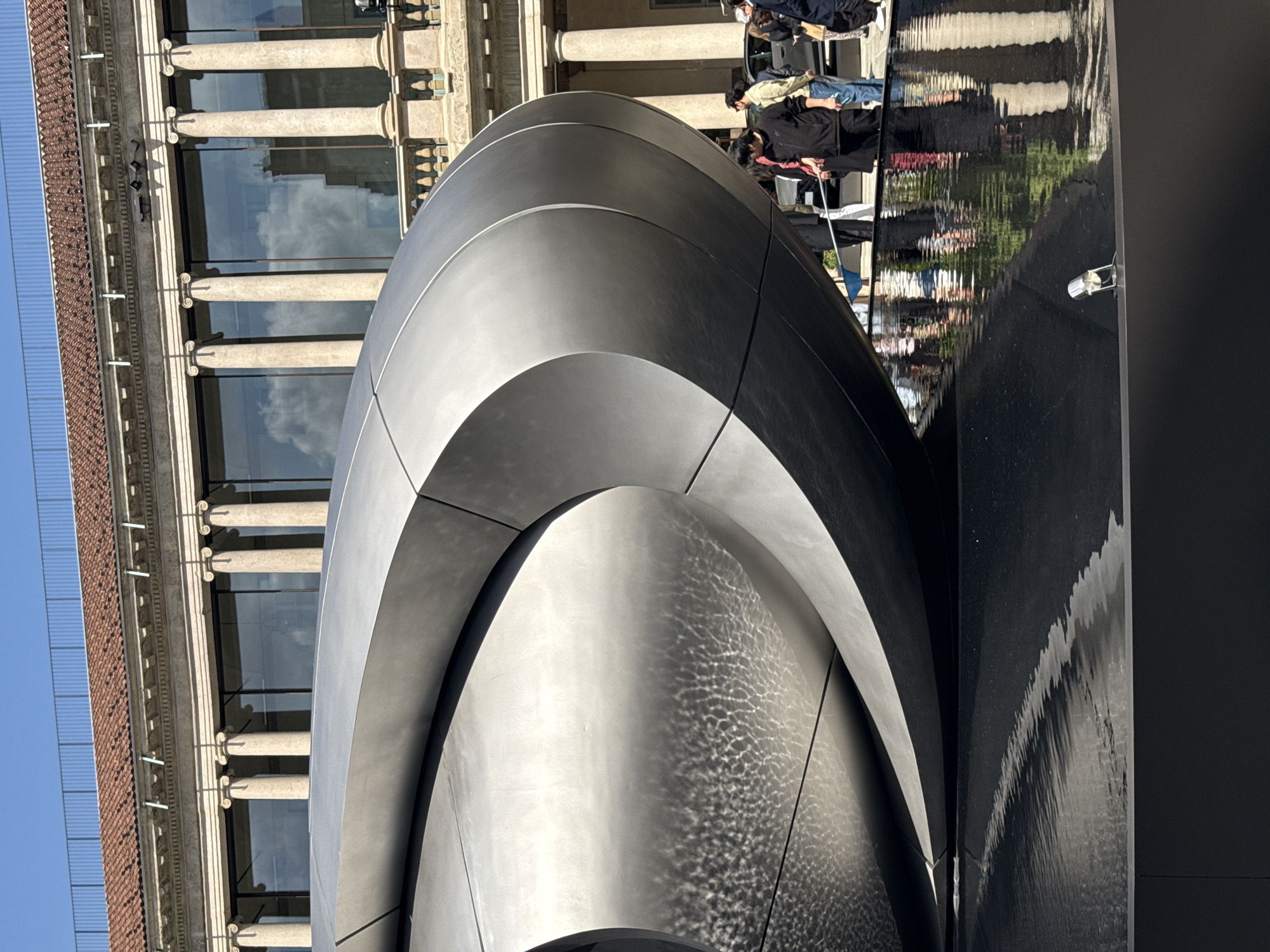 The image, taken in Milan, Italy, presents a dynamic contrast between modern design and classical architecture. In the foreground, a large, imposing sculpture or architectural element dominates the right side. It is crafted from a dark, possibly brushed metal, featuring smooth, curvilinear surfaces that form prominent arch-like structures. Sunlight highlights its contours, and intricate, ripple-like patterns, originating from a nearby water feature, dance across its lower sections.
To the left, an ornate, multi-story classical building stands, adorned with numerous columns featuring decorative capitals. Its large windows reflect a bright, clear blue sky and scattered clouds. The building's roof is covered with reddish-brown tiles, and its facade is rich with detailed cornices and architectural embellishments.
On the far right, a tall, dark, and highly reflective surface, likely a sheet of falling water or a calm pool, captures distorted reflections of people and the surrounding environment. The reflections appear stretched and rippled, suggesting movement. Several individuals are visible in the mid-ground, standing near this reflective surface and partially obscured by the modern sculpture. They are casually dressed, with some wearing face masks, appearing to be observing the scene or passing through the area.
The scene unfolds under bright daylight, indicating a clear and sunny day. The blend of the sleek, futuristic lines of the metallic structure and the intricate, historical details of the classical building creates a visually engaging composition. No discernible text is visible within the image.