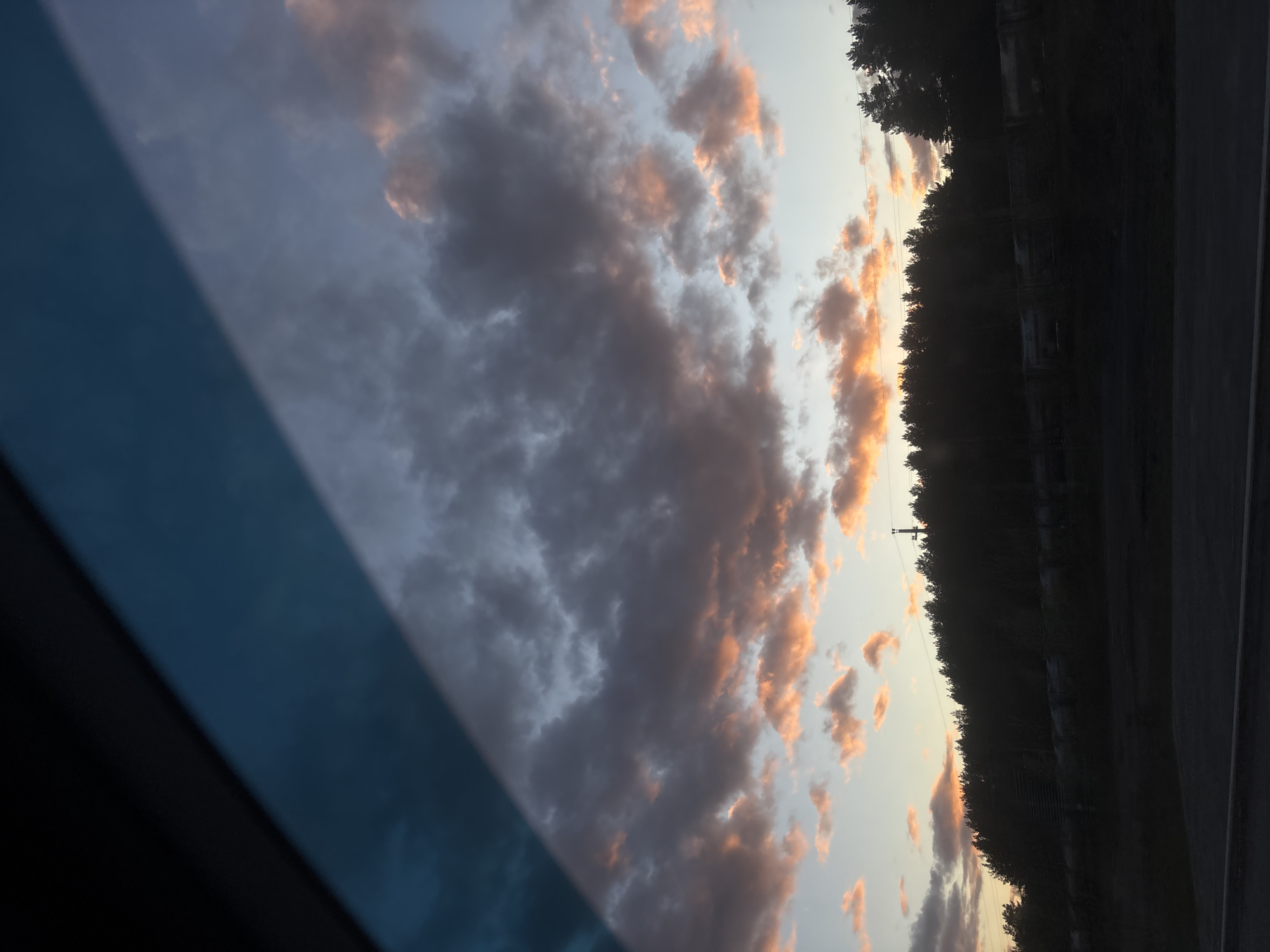 The image depicts a dusk or dawn sky with orange-tinted clouds, partially obscured by a dark blue, possibly automotive, interior element on the left. A line of trees borders the sky on the right, with a telephone pole barely visible amidst them. Adjacent to the trees is a dark, reflective surface, likely asphalt or road, with a light dividing line suggesting a road. The image is not sufficient to determine its specific location in Perm, Russia.