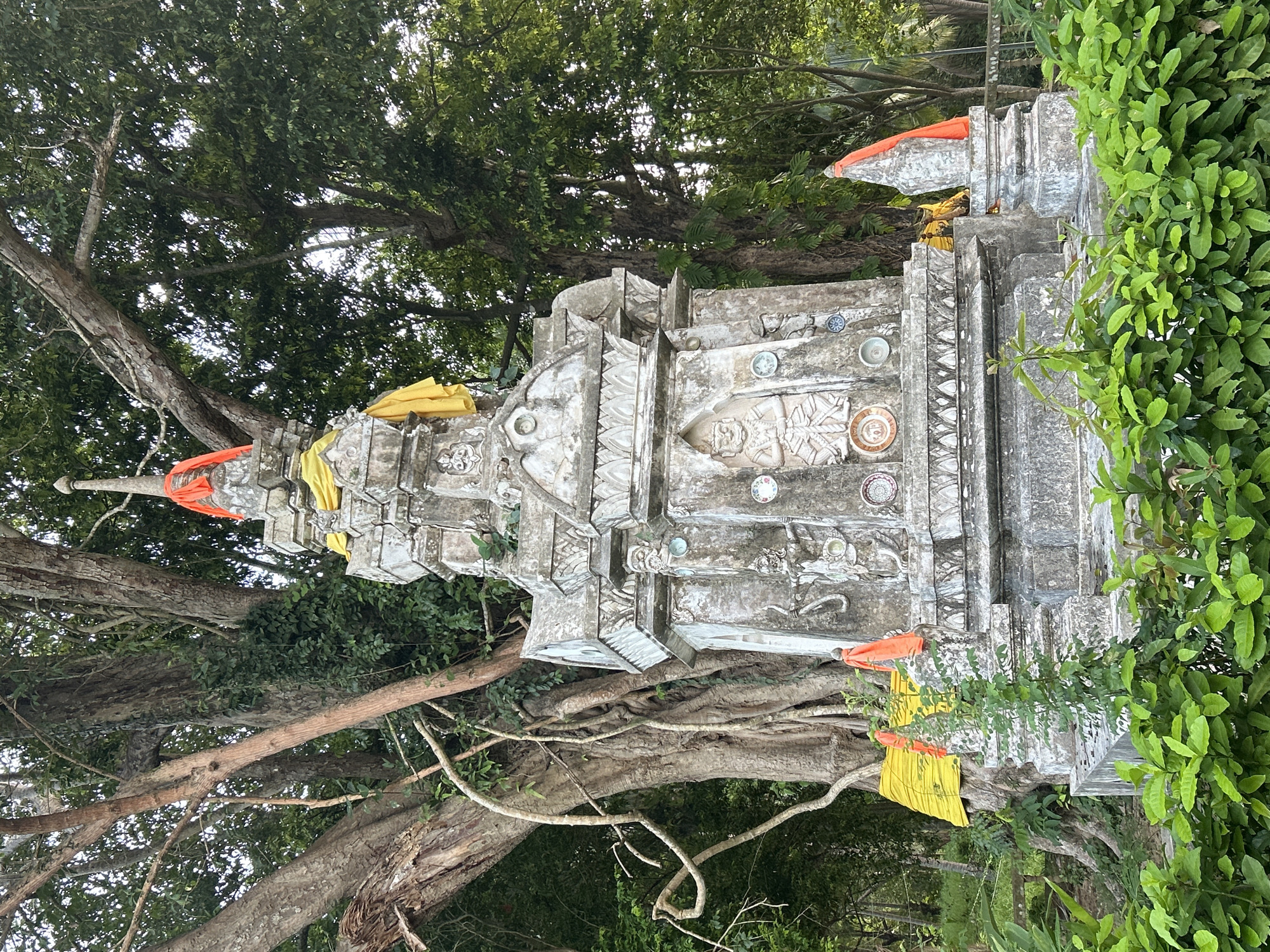 A full shot captures a weathered, ornate stone structure adorned with orange and yellow fabric, set against a backdrop of lush green foliage and the massive trunk of a banyan tree. The structure, likely a stupa or shrine, features intricate carvings, including a central figure of a warrior or deity, and is decorated with various small ceramic plates. The banyan tree's roots are visible, entwined with the shrine, suggesting a long-standing presence in its environment. The scene appears to be outdoors in a tropical setting, possibly a temple ground or a significant natural landmark within the Ko Pha-ngan District, Thailand, indicated by the surrounding vegetation and the style of the structure. There are no people visible in the frame. The lighting suggests it is daytime, with soft sunlight filtering through the leaves.