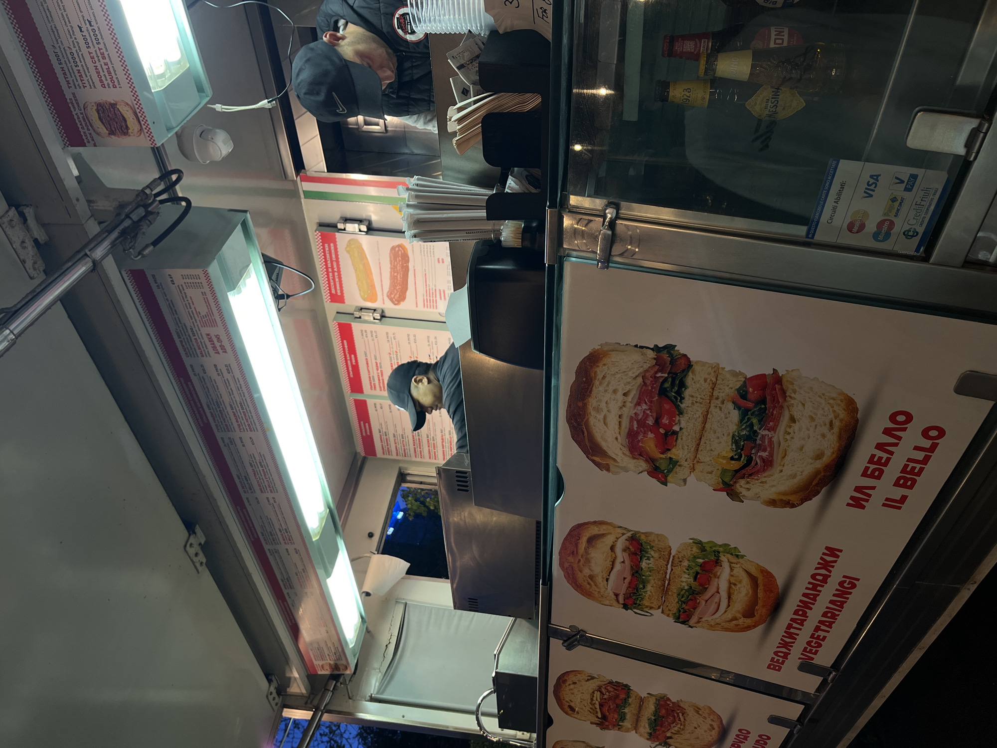This media file captures a nighttime scene inside a food truck or kiosk in Sofia, Bulgaria. A vendor, wearing a black cap and jacket, is seen leaning over a stainless steel counter, possibly attending to a customer or preparing an order.
The interior of the kiosk is brightly lit by several fluorescent light fixtures. On the right, a large vertical poster showcases appealing images of various sandwiches, accompanied by text in both Bulgarian Cyrillic and Italian, such as "ВЕДЖИТАРИАНДЖИ VEGETARIANGI" and "ИЛ БЕЛЛО IL BELLO." Below the counter, a refrigerated display case holds several beverage bottles, including "PERONI," "BIRRA MESSINA," "ODINO," and "ONI." Affixed to the glass is a sign detailing accepted payment methods, listing "Mastercard," "VISA," "Maestro," "PagoBANCOMAT," "VPay," and "CrediFriuli," with the Italian phrase "IN QUESTO ESERCIZIO LE CARTE SONO ACCETTATE" (In this establishment, cards are accepted).
Along the top and left side of the kiosk, menu boards display food items with descriptions and prices, featuring both Bulgarian and English text like "BLACK DOUBLE CHEESE," "TITAN 500G," and "DOUBLE SET 500G." Dispensers for napkins and straws are visible on the counter. The overall setting indicates a street food business operating during the evening hours.