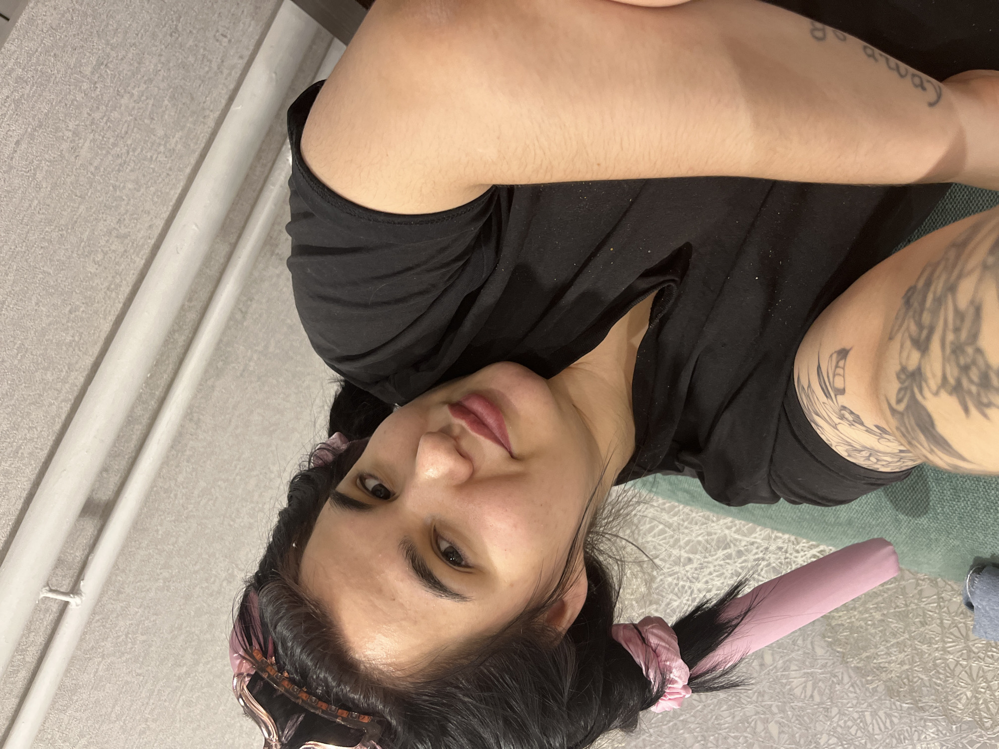 A young adult woman with dark hair is lying on her side, wearing a black t-shirt. Her hair is styled with pink hair accessories. She has tattoos on her left arm. The location is not discernible from the provided image, and there is no indication that it is in Chaikovsky, Russia. The background shows a wall and a textured surface.