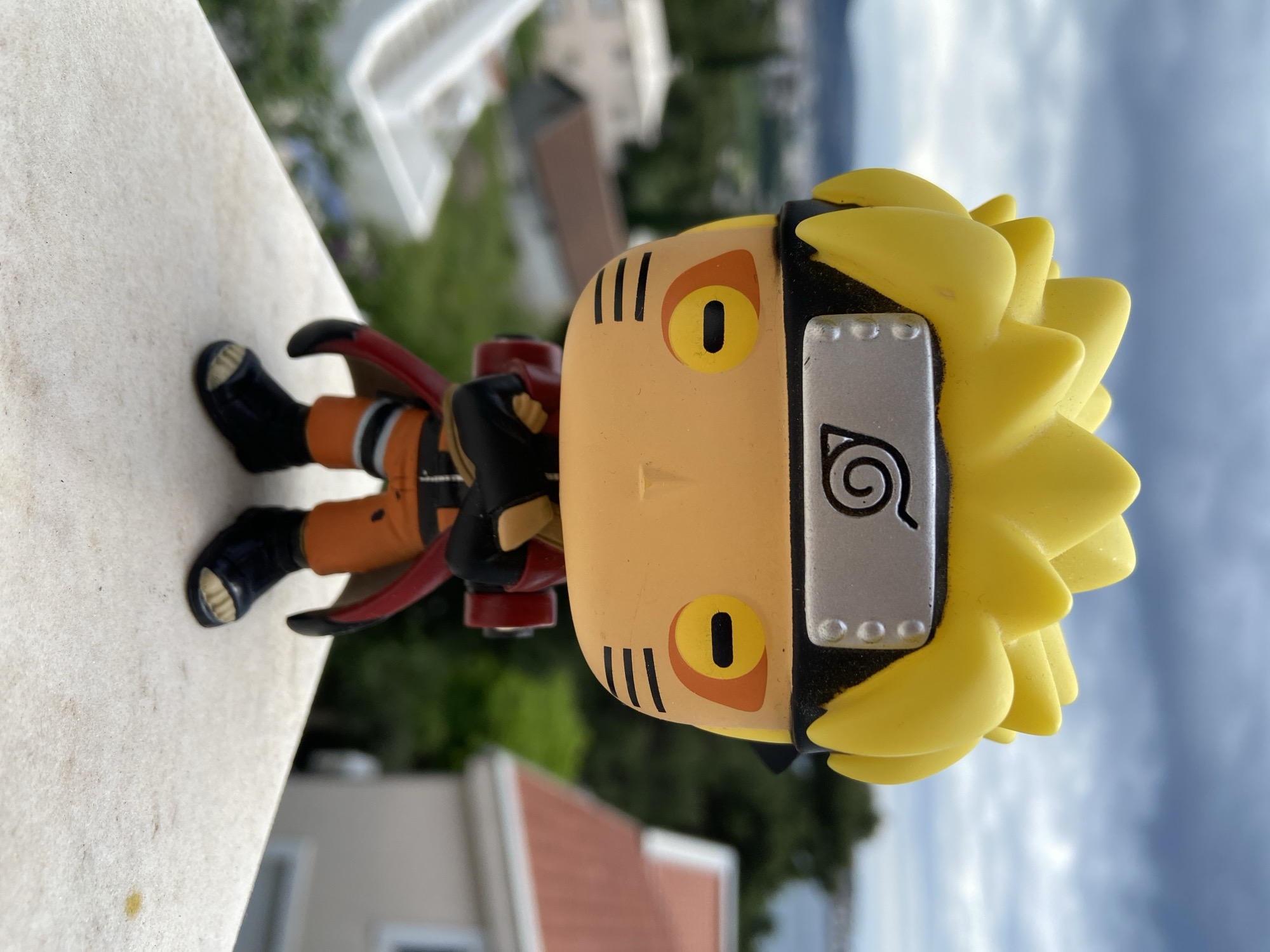The image features a Naruto Funko Pop figurine, photographed outdoors in Šušanj, Montenegro. The figurine is positioned on a white ledge or railing, with a blurred background showing buildings and a distant view of the sky and landscape, typical of the Montenegrin coast. The focus is sharp on the figurine, detailing its features such as the yellow spiky hair, the Hidden Leaf Village headband, and the orange jumpsuit. The figure’s black shoes are visible on the left edge of the frame. The overall setting suggests a residential area during daylight hours.