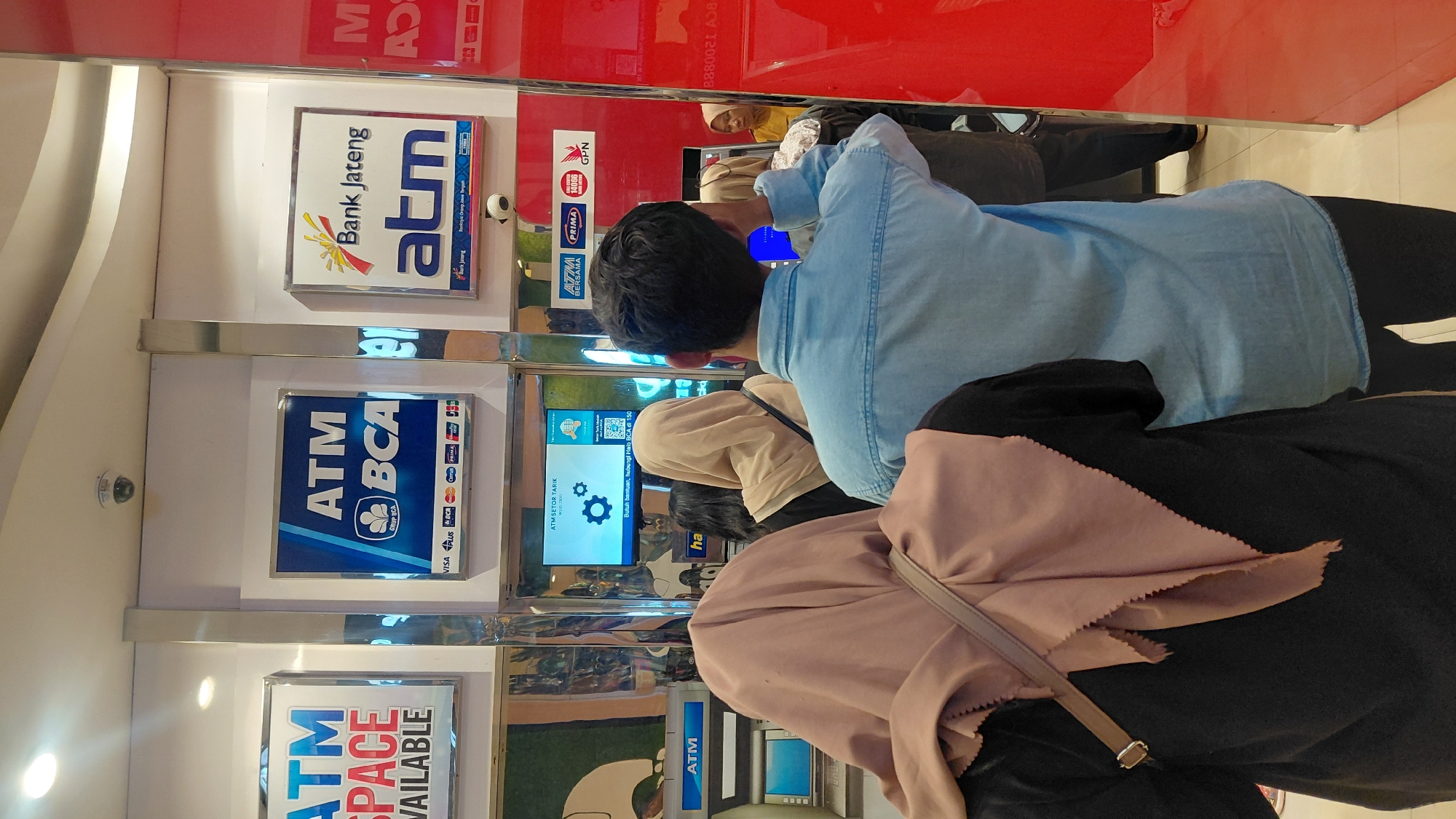 The image shows an ATM lobby in Purwokerto, Indonesia. Several people are standing in line to use the ATMs. One ATM is visible on the left, displaying a blue screen. Above the ATMs, signs indicate "ATM SPACE AVAILABLE," "ATM BCA," and "Bank Jateng atm." A security camera is mounted on the ceiling. The environment appears to be indoors, likely a bank or a shopping mall. The lighting is bright, illuminating the scene. The people in the image are dressed casually, and most of them are wearing hijabs. The overall atmosphere seems to be one of a typical banking transaction.