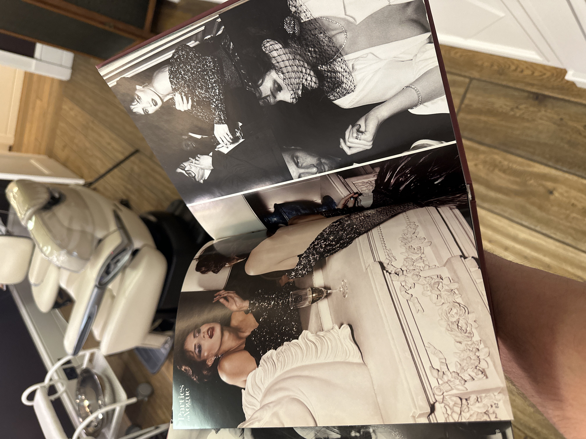The image shows an open book, "Parties in Vogue", displaying black and white photographs. One page features a woman in a sequined dress, reclining near a fireplace with a champagne glass. The opposite page depicts a gathering of people, including a woman in a veiled hat and a man with glasses. The photographs suggest a glamorous, high-fashion social event. The image is held by a person whose arm is visible. In the background, there's a massage chair and wooden flooring. The location is not discernible from the image provided. Moscow, Russia cannot be determined from the image.