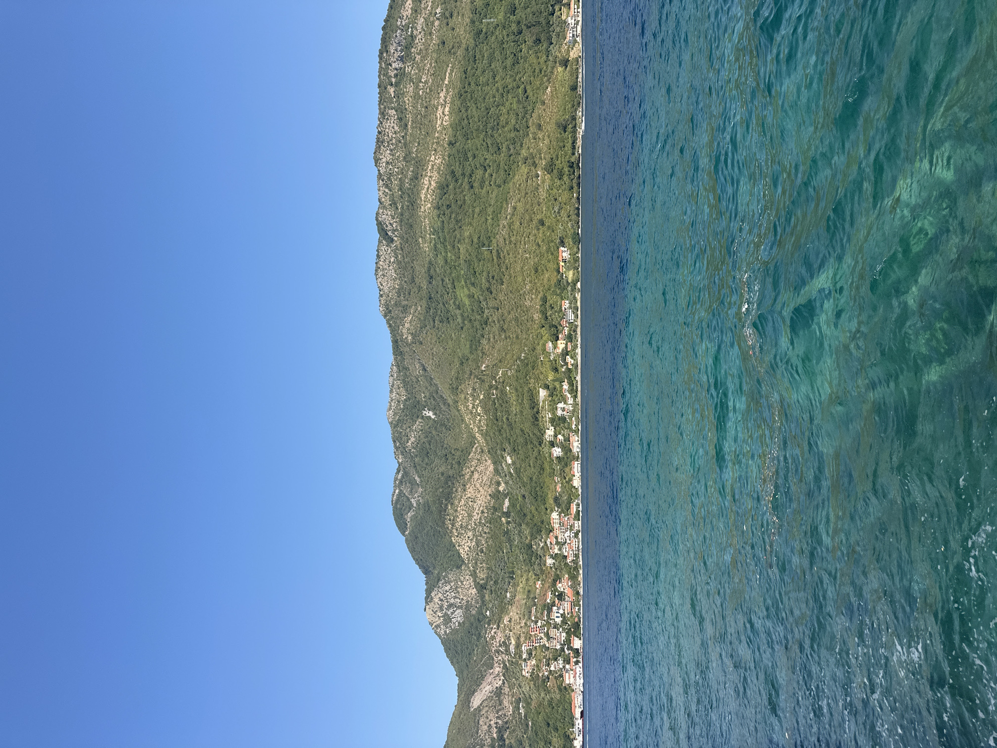 The image depicts a view of the Bay of Kotor from the water, likely taken from a boat. In the foreground, the clear, turquoise water is visible with ripples and some foam on the surface. The middle ground shows a coastline with lush green hills rising behind it. Along the shoreline, there is a settlement with numerous buildings featuring red-tiled roofs, characteristic of the architecture in the region. The buildings are clustered together and extend up the gentle slope of the hill. In the background, a large, verdant mountain range dominates the landscape, with some rocky outcrops visible. The sky is a clear, vibrant blue, indicating a sunny day. A few boats can be seen on the water near the shore, and power lines are visible ascending the hills. The scene suggests a peaceful coastal environment in a town like Lepetane, Montenegro, known for its scenic beauty and position within the Bay of Kotor.