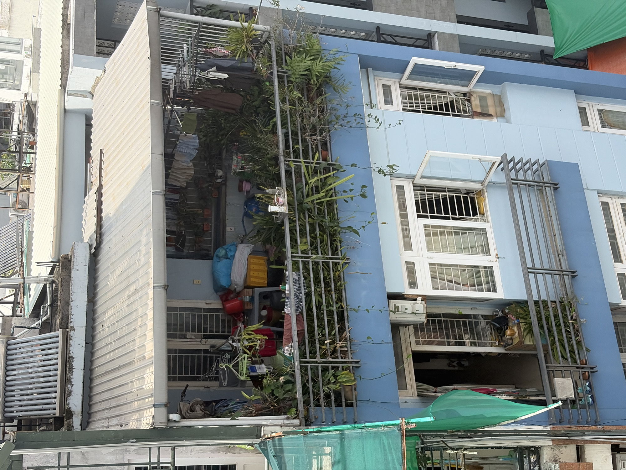 The image, captured from an elevated perspective and oriented on its side, depicts a multi-story residential building and its immediate surroundings in an urban area, consistent with the location context of Thủ Đức, Vietnam.
The central subject is a residential structure painted in varying shades of light blue, featuring multiple windows and balconies across its visible floors. Many windows are open and fitted with white metal security grilles. Abundant greenery, including ferns and climbing plants, grows on balconies and along vertical metal frameworks attached to the building's facade, indicating an integration of plant life into the urban dwelling.
Daily life activities are evident, with clothes hanging to dry on several balconies. These outdoor spaces also serve as storage areas for various household items, such as blue and yellow bags, red buckets, and other containers. A bicycle and what appears to be a fan or small appliance are also visible on a balcony. Air conditioning units are mounted below some windows.
To the left of the blue building, a structure with light-colored, corrugated metal sheeting forms a wall or an extension, with a large metal grille or gate adjacent to it. In the lower foreground, a green tarp or awning provides shade.
The scene is brightly lit by natural sunlight, indicating daytime and clear weather. A small, dark number, possibly "24" or "27", is faintly discernible on a white electrical outlet on the wall of one of the balconies. The overall impression is of a lived-in, practical, and densely packed urban residential environment.