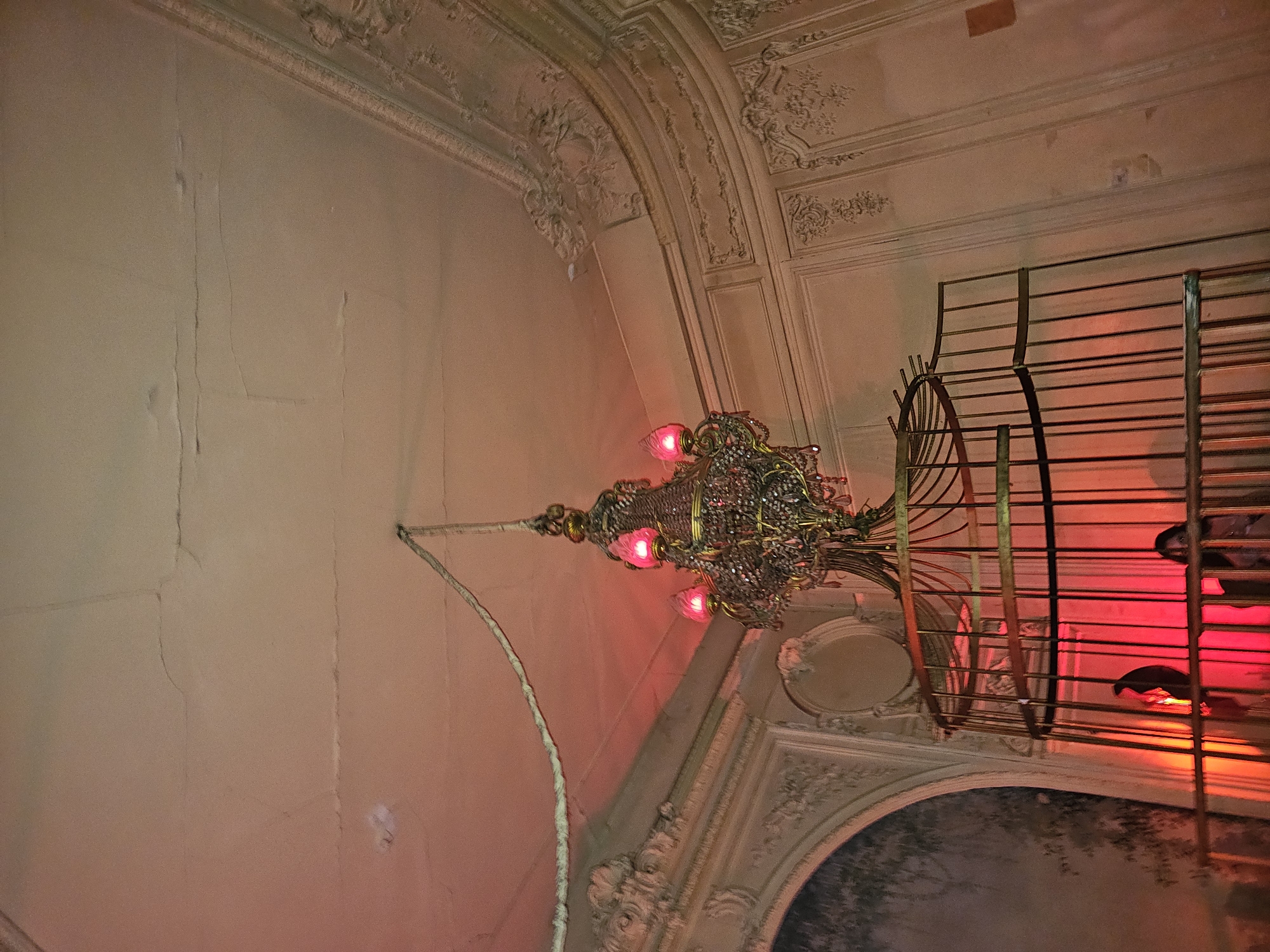 The image shows a chandelier hanging from the ceiling of a building in Paris, France. The walls are decorated with elaborate plaster moldings. There are cracks in the walls. There is a metal structure next to the chandelier with a person standing in the structure. The light in the building is red.