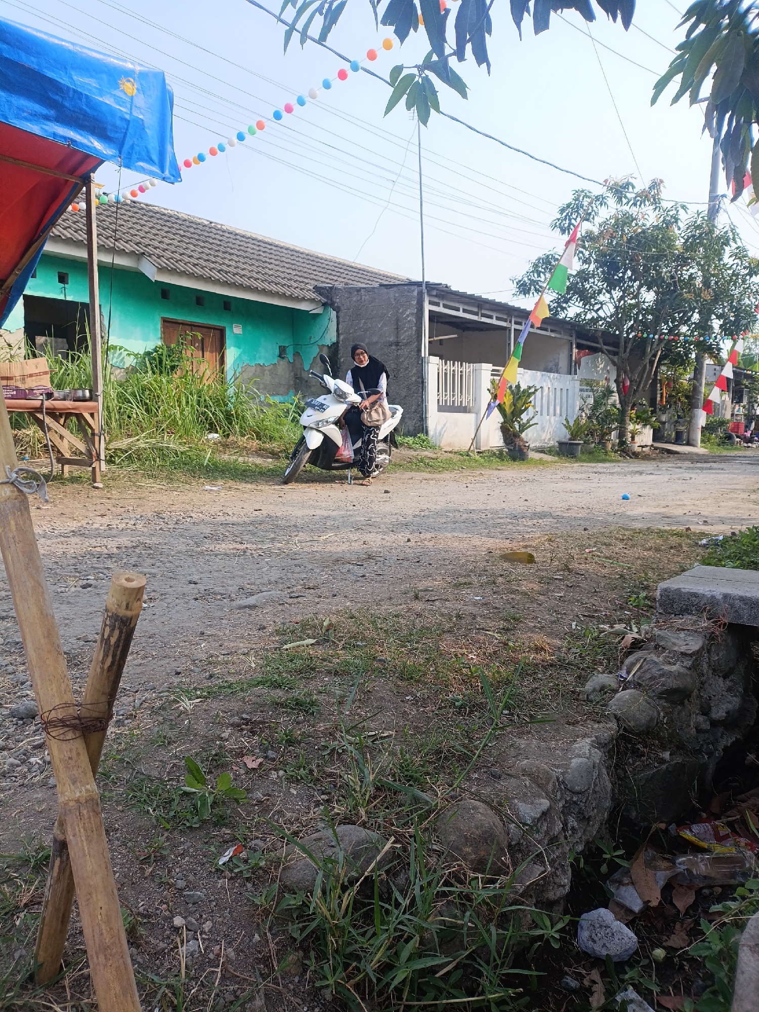 In Cariu, Indonesia, a woman wearing a black hijab and patterned pants sits on a white scooter parked on a dirt road. A brown shoulder bag hangs from the scooter's handlebars. To the left of the scooter, a bright teal-colored house with a gray roof is visible, partially obscured by tall green grass and a red and blue tarp supported by bamboo poles. Above the house, strings of colorful spherical decorations are strung across the scene, with some colorful flags also visible on poles further down the road. A tree with green foliage stands to the right, adorned with more flags. The ground is uneven, with scattered rocks and dry vegetation. In the foreground, two bamboo poles are tied together with wire. A small, dark stone structure with rocks and debris is situated to the lower right.