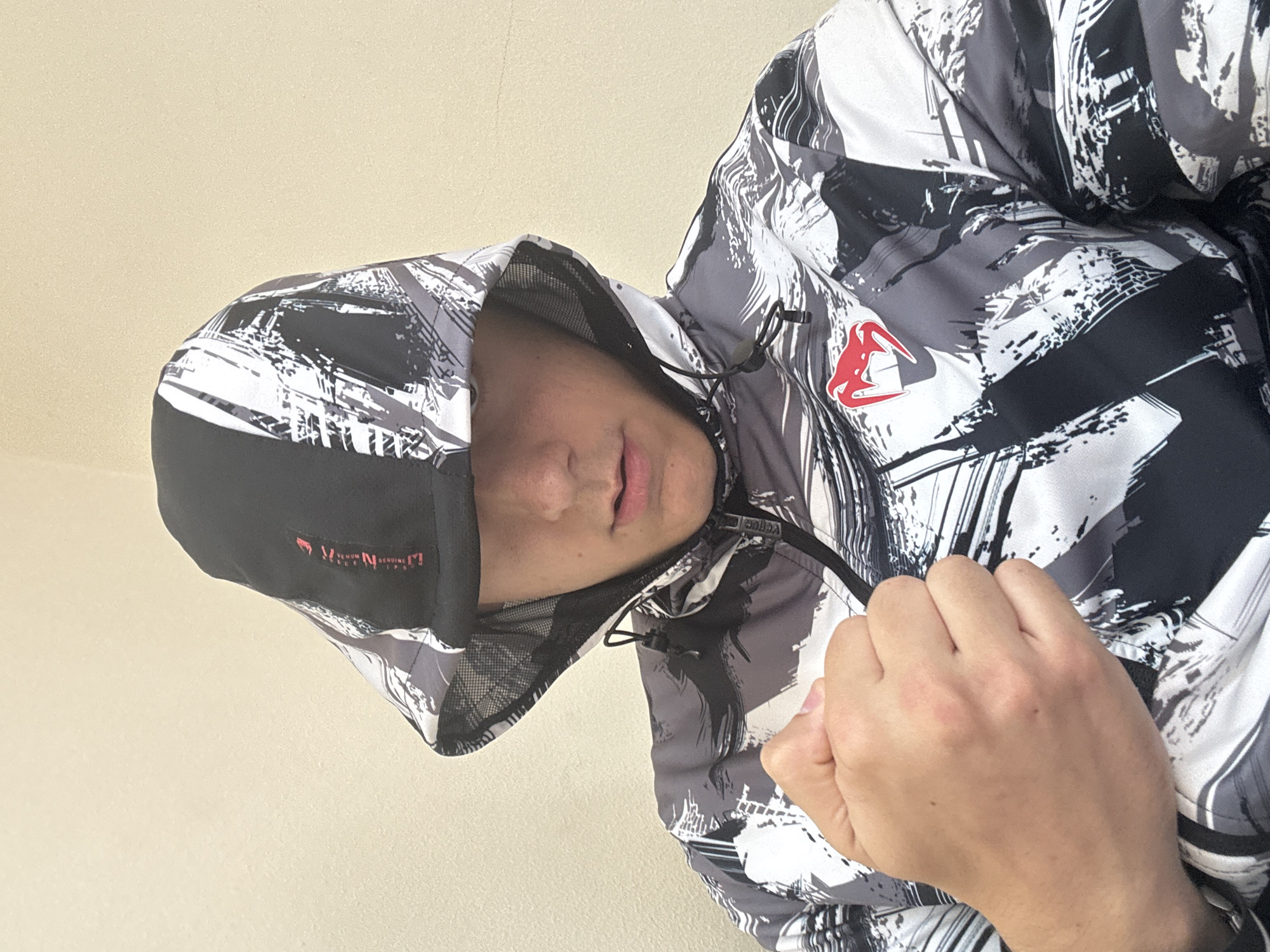 A tilted, close-up shot captures a person, likely male, wearing a distinctive hooded jacket against a plain, light beige background. The person's face is partially visible within the hood, showing their chin, mouth, and nose, with a neutral expression. Their right hand is clenched into a fist, resting on the jacket near the central zipper.
The jacket features a bold, abstract pattern in black, white, and various shades of gray, resembling distressed paint strokes or urban camouflage. A prominent red logo, stylized as an animal head or claw mark, is visible on the left chest area of the jacket. On the black band at the front of the hood, red text reads "VENUM GENUINE" and partially "SPORT" or "SPORTS," identifying the brand. The interior of the hood appears to have a mesh lining.
The lighting is soft and even, suggesting daytime conditions, possibly indoors or on an overcast day, as there are no harsh shadows. The plain background offers no specific location cues, but the context indicates the image was taken in Plovdiv, Bulgaria. The person is posed, highlighting the design and branding of the jacket, which suggests suitability for cooler weather or an athletic setting.