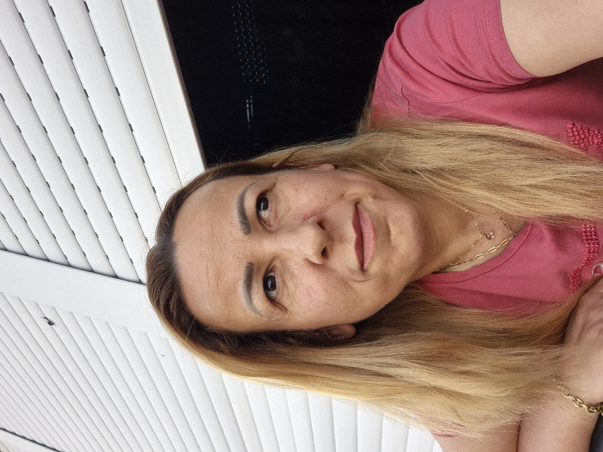 A woman with long blonde hair and a pink t-shirt is looking off to the right of the frame. She is wearing a gold necklace with a small pendant and a gold bracelet. The background is a white corrugated metal wall. The lighting is natural and appears to be from the side.