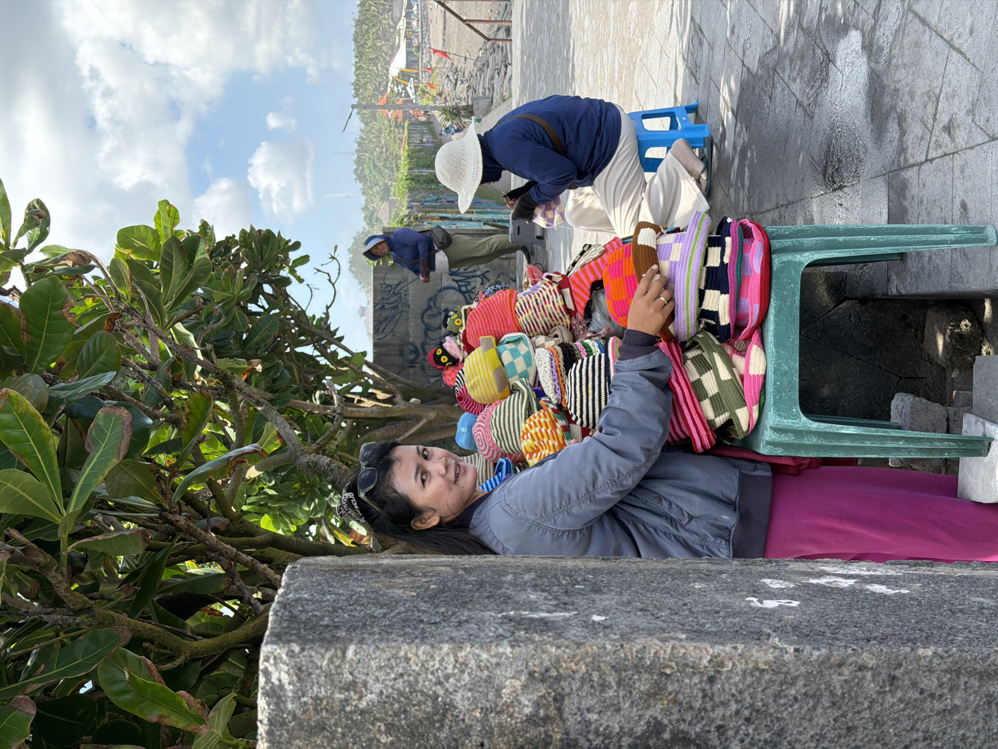 The image depicts a street scene in Mengwi, Indonesia, near a beach. In the foreground, a woman is standing behind a table laden with colorful, handmade crocheted hats. She is wearing a gray jacket and pink pants, and is looking towards the camera with a slight smile. To her right, another woman, wearing a blue top and a white hat, is seated on a blue stool, crocheting. In the background, a third person in a blue hooded jacket stands near a wall. The environment suggests a marketplace or stall setup, with a beach and palm trees visible in the distance. The ground is paved with stone, and the sky is partly cloudy.