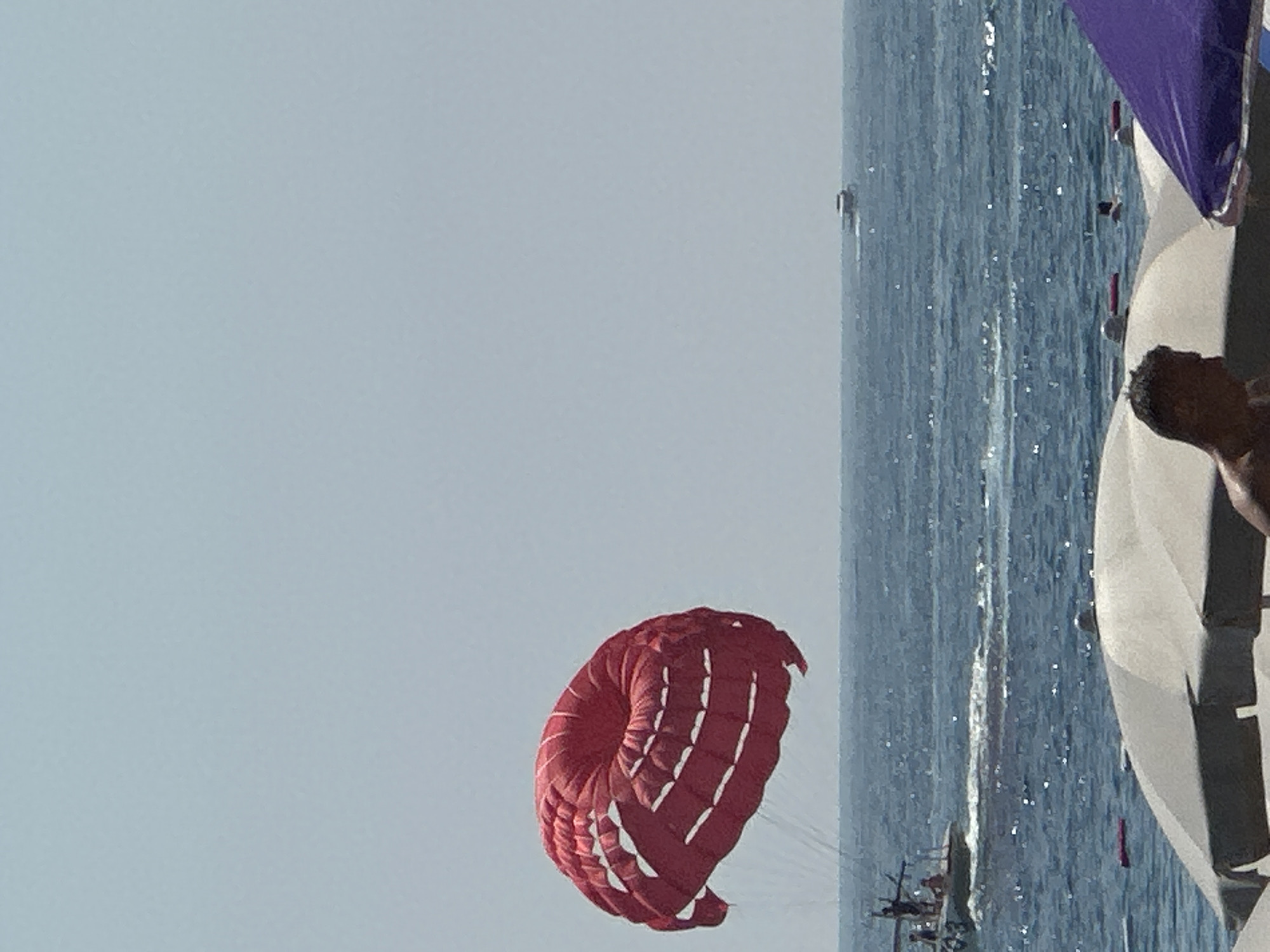 A red parasail is suspended in the sky above a blue, choppy ocean. The parasail appears to be attached to a boat in the water, which is visible in the lower left corner of the image. The boat has several people on it. In the distance, another smaller boat is visible on the horizon. The foreground of the image shows the tops of beach umbrellas, with a person's head visible in the lower right corner, suggesting a beach scene in Budva, Montenegro. The sky is clear and a light blue color.