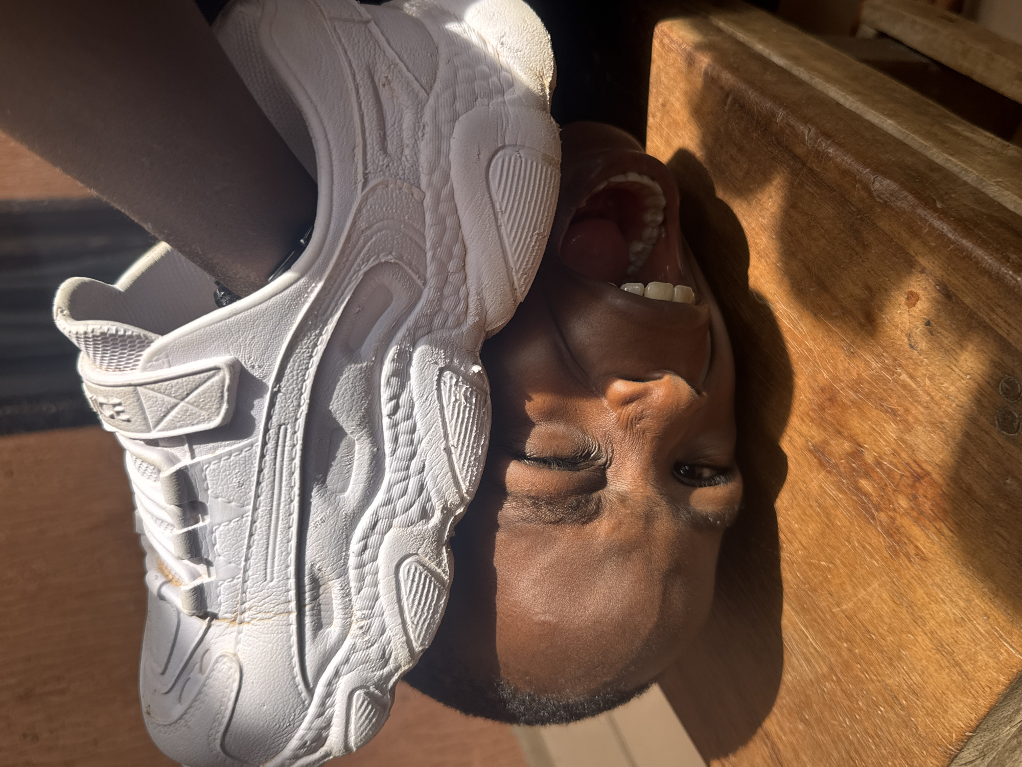 A child is upside down with their foot crammed into the opening of a white sneaker. The child's mouth is open, exposing their teeth. The scene is illuminated by sunlight, casting shadows on a wooden surface. No details indicate a location in Onitsha, Nigeria.